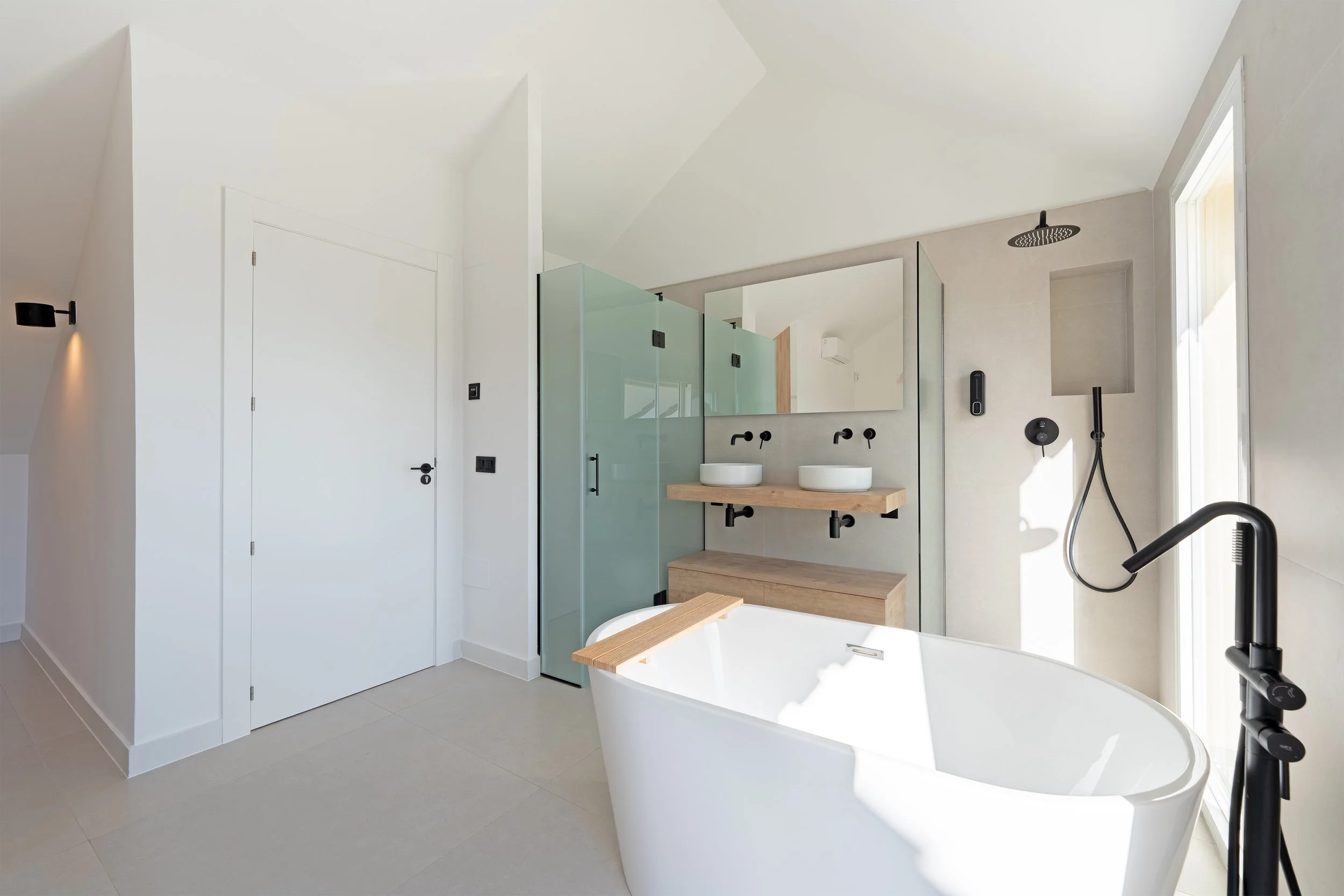 Modern bathroom with a freestanding bathtub, double sink vanity with large mirror, glass shower enclosure, black fixtures, and minimal decor, bright natural light from window.