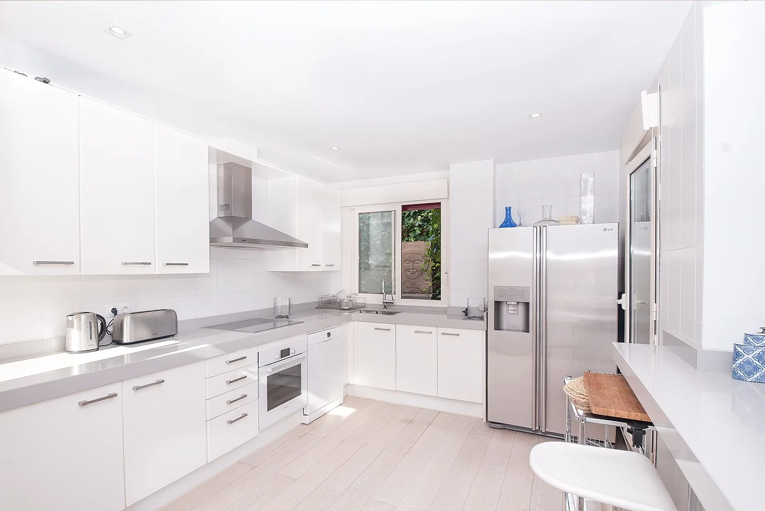 Modern white kitchen with stainless steel appliances, a window with greenery outside, and kitchen accessories on the counters.