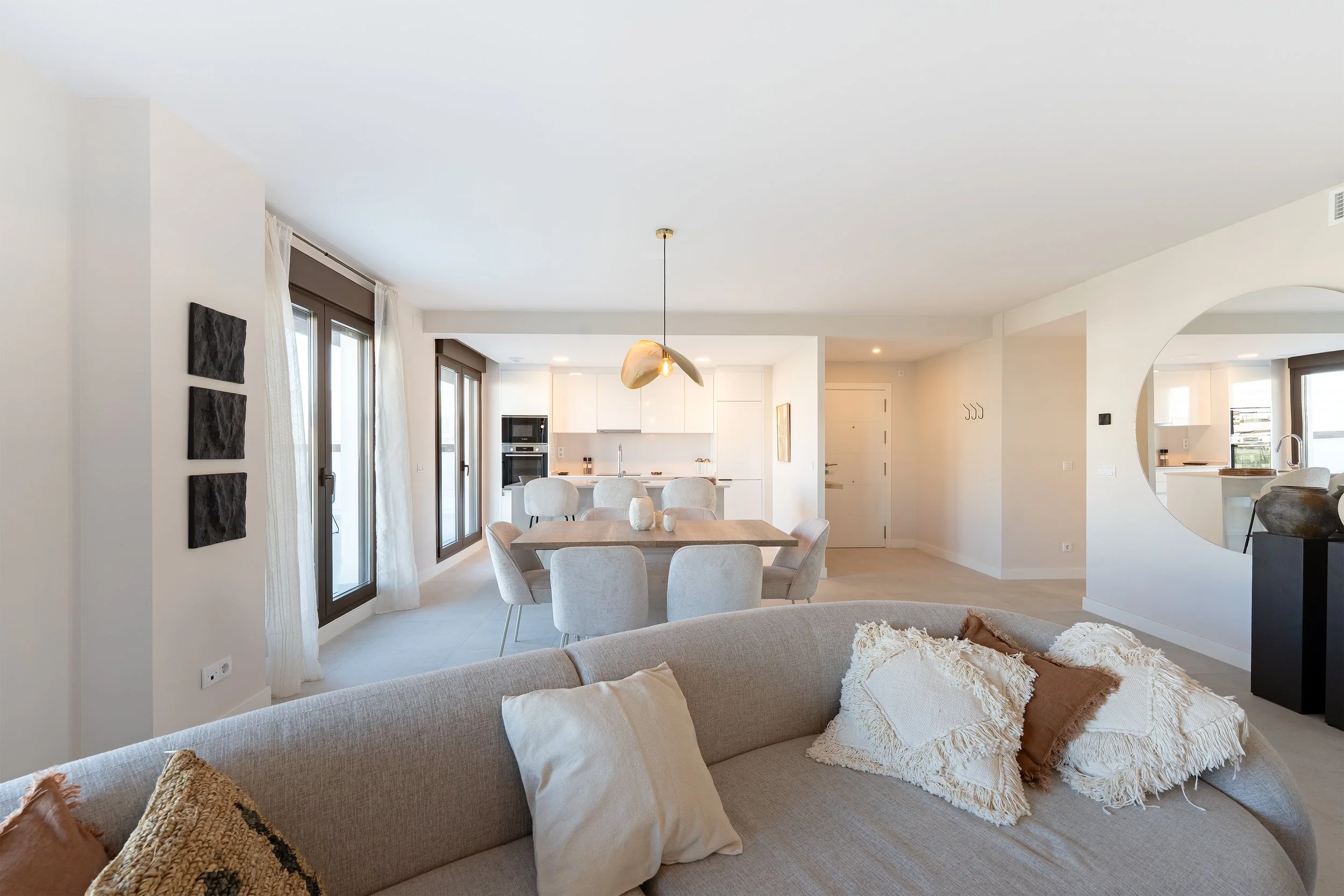 Open-concept living and dining area with white walls, large windows, a round mirror, beige sofa with pillows, dining table with six chairs, and kitchen in the background.