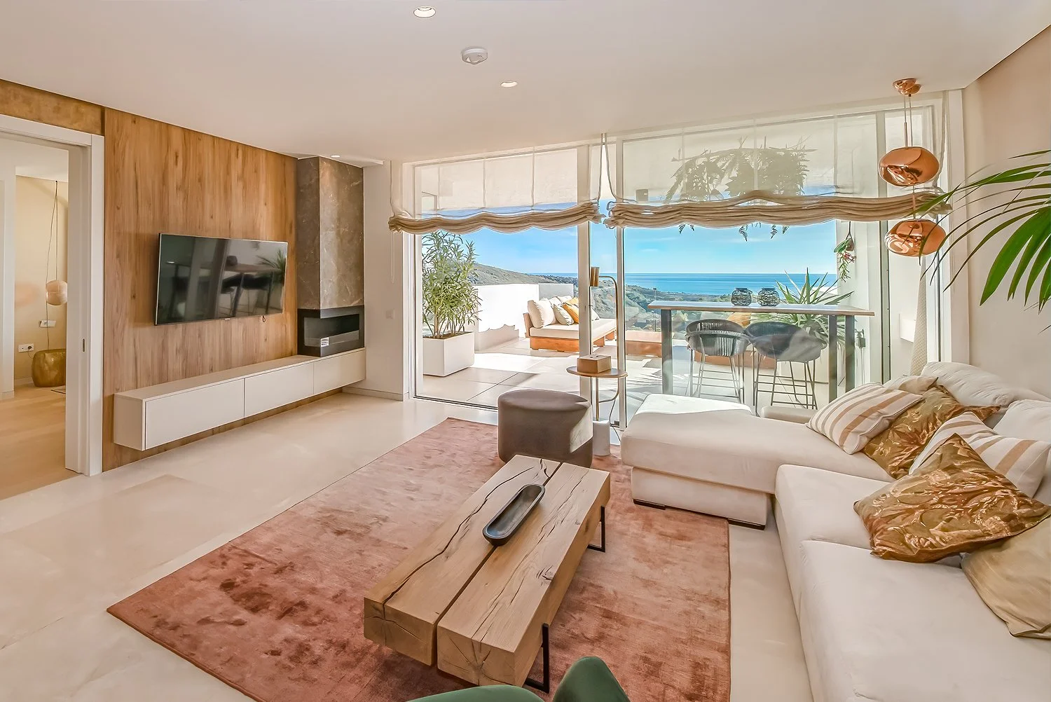 Living room with sofa, coffee table, mountain landscape and ocean view through large glass doors, and balcony with outdoor seating.