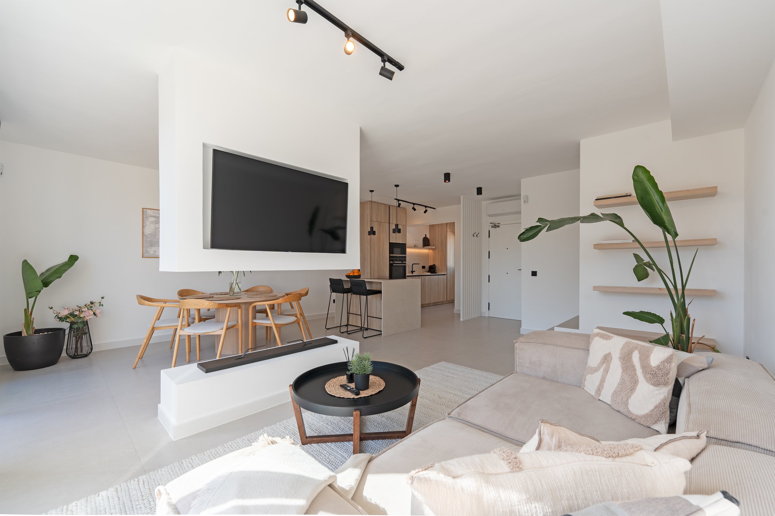 Modern open-plan living room and kitchen with white walls, large flat-screen TV, beige sofa with patterned pillows, black round coffee table with small potted plant, wooden dining table with chairs, and kitchen with light wood cabinets, black applian
