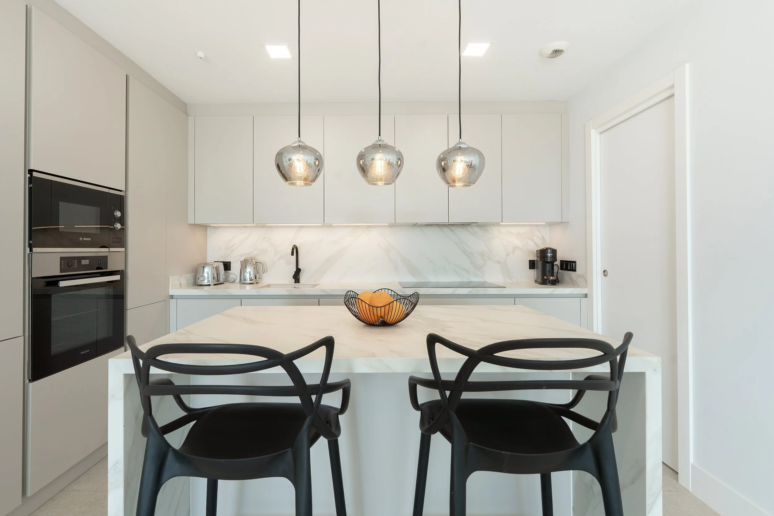 Modern kitchen with white cabinets, marble countertops, and black chairs around a marble island with a fruit bowl. Three metallic pendant lights hang above the island.