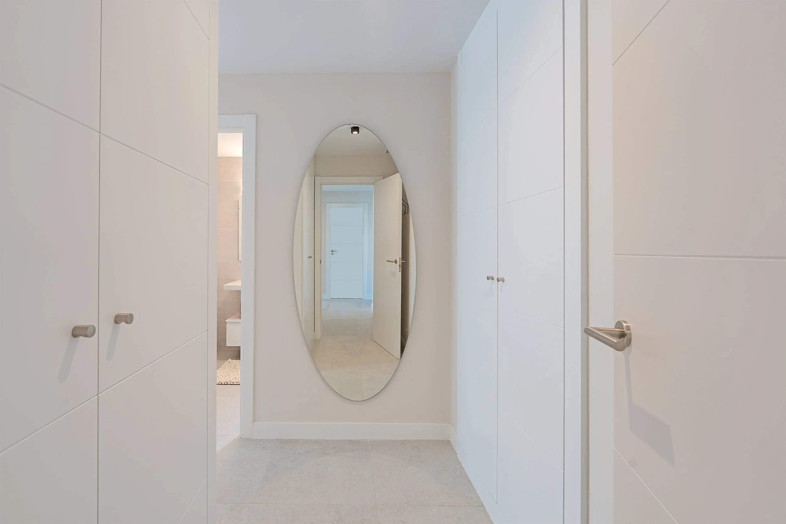 A hallway with white built-in closets on either side, a full-length oval mirror on the back wall, and a view of a white door at the end of the corridor.