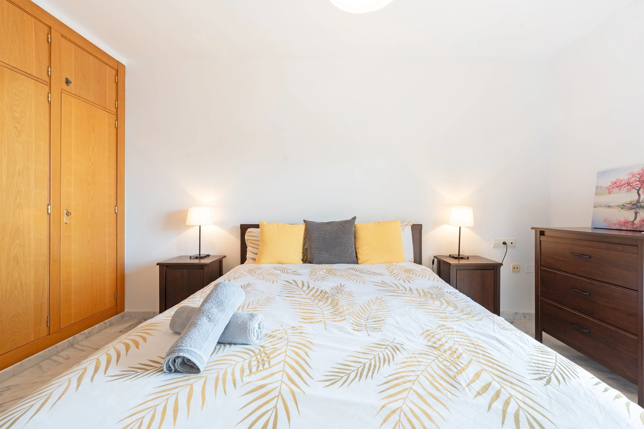 A bedroom with a bed covered in a white and gold leaf patterned bedspread, four pillows in yellow and gray, two rolled towels on the bed, wooden nightstands with lamps on each side, a wooden dresser with a pink floral painting on top, and a large woo