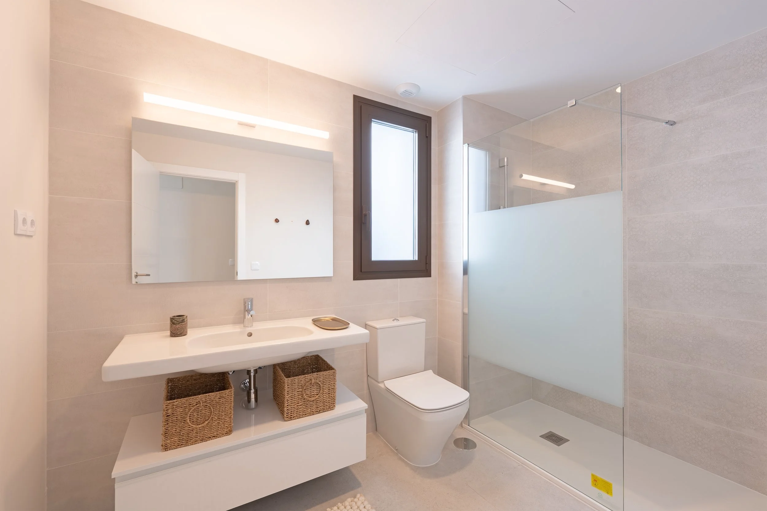 Modern bathroom with beige tile walls, large mirror, wall-mounted sink, wicker baskets, white toilet, frosted glass shower enclosure, and a window with a dark frame.
