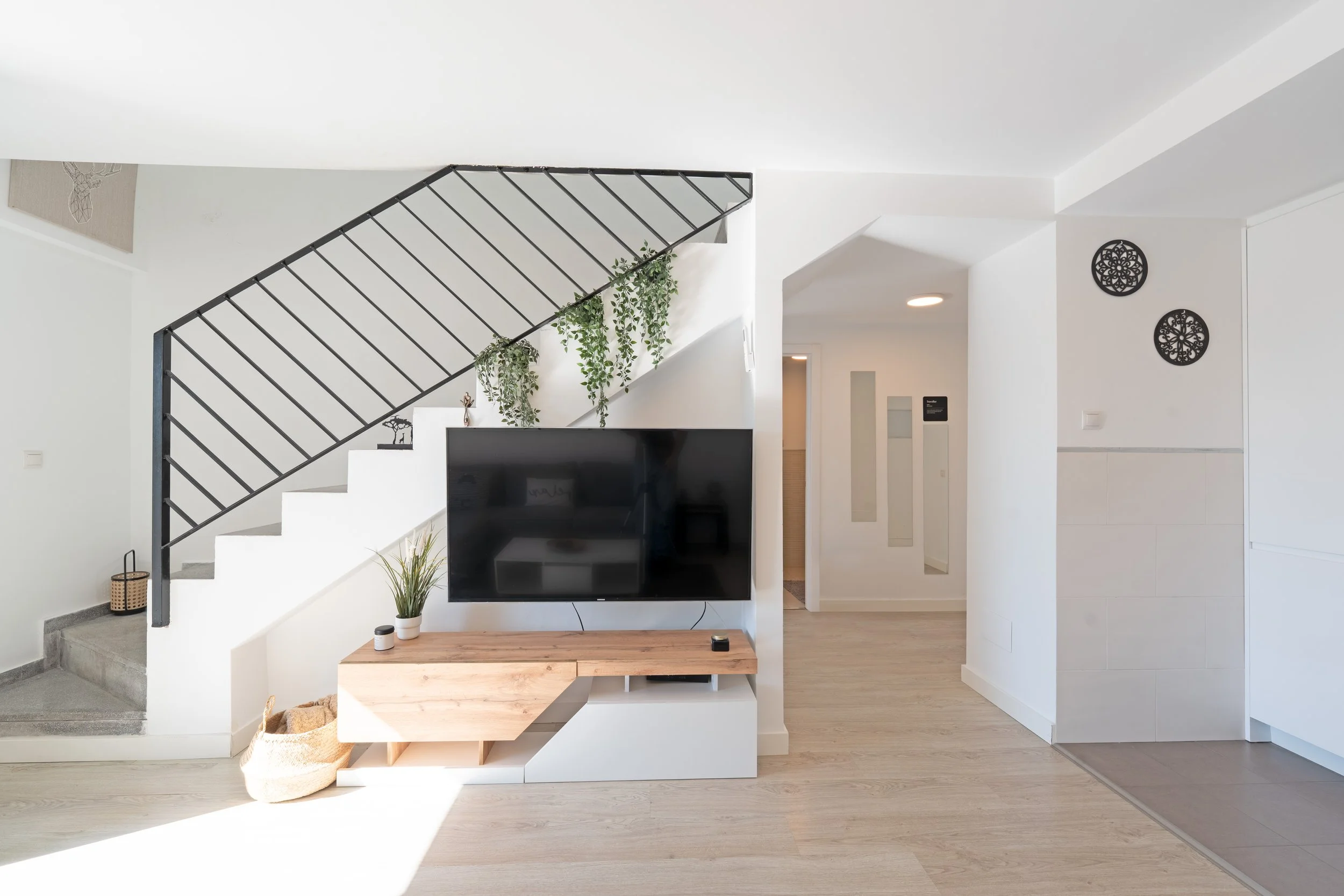 Modern living room with white walls, a staircase with black railing, a wall-mounted flat-screen TV, wooden console, decorative plants, and black wall art.