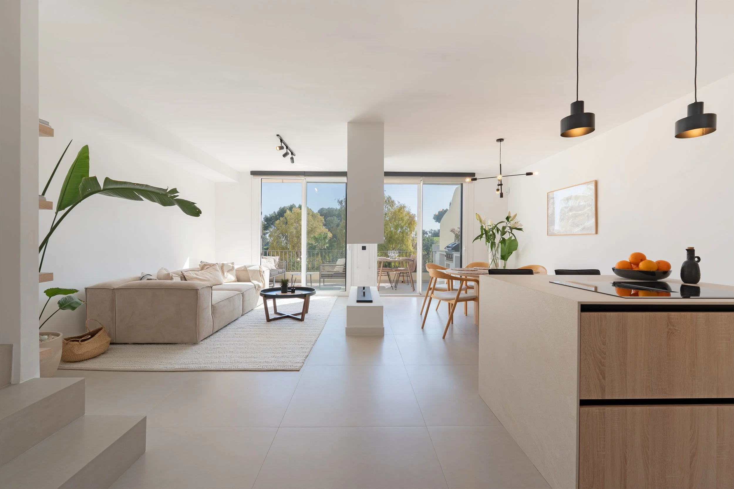Modern, bright living room and kitchen with large sliding glass doors opening to a balcony with trees outside. The space features a beige sofa, a round coffee table, a dining table with chairs, black and wooden light fixtures, greenery, and minimalis