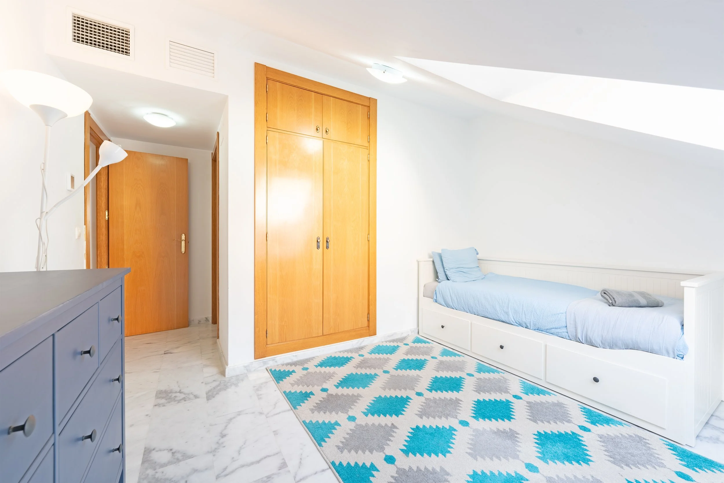 A bedroom with a bed in white frame, blue bedding, pillows, a built-in closet with wooden doors, a blue dresser, and a patterned rug on a marble floor, illuminated by ceiling lights and a skylight.