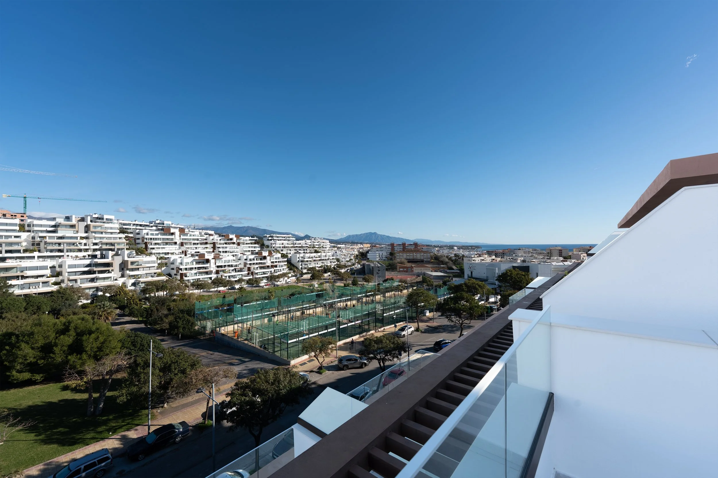 View from a balcony overlooking a cityscape with white apartment buildings, tennis courts, parking lots, trees, and mountains in the distance under a clear blue sky.