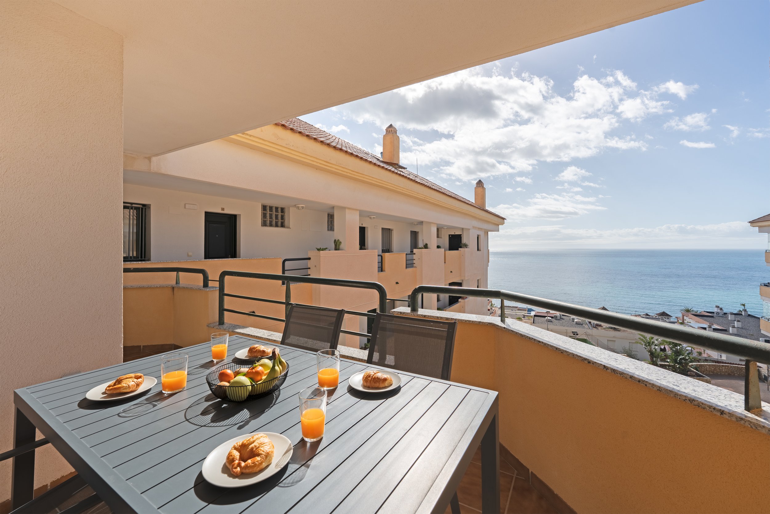 Balcony with a table set for breakfast, including croissants, orange juice, and fresh fruit, overlooking the ocean on a sunny day.