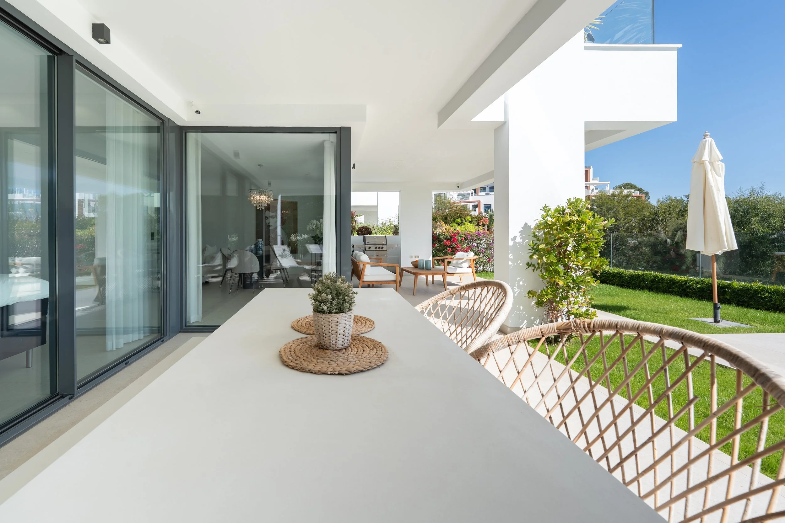 Outdoor patio with a large white table, woven chairs, potted plant, and lush green lawn; glass doors lead to interior dining area.
