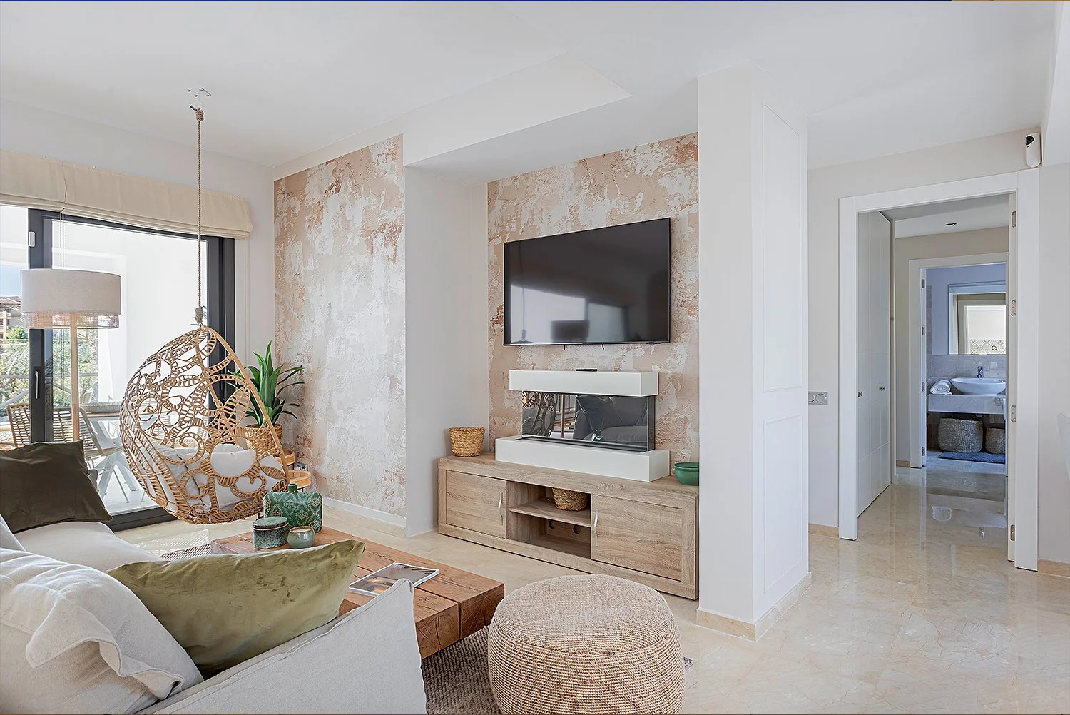 Living room with beige tiled floor, a cream sofa with pillows, a wooden coffee table, a hanging wicker chair with a cushion, sliding glass door to balcony, a wall-mounted TV above a modern fireplace, textured wall behind TV, white doors leading to ba