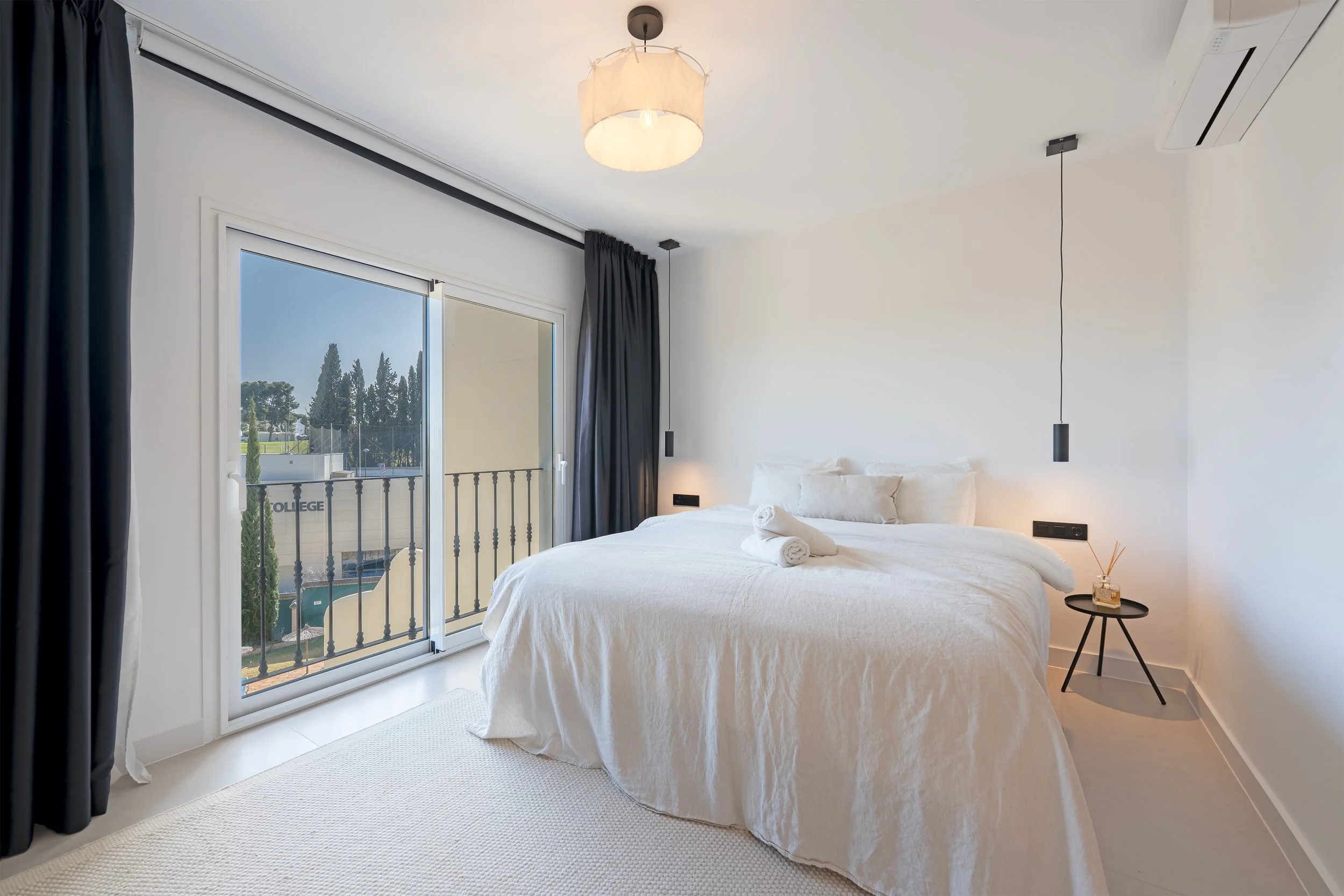 A modern bedroom with a white bed, dark curtains, a sliding glass door leading to a balcony, and minimalist black bedside table with a diffuser and reed sticks, featuring a simple ceiling light and wall-mounted pendant light.