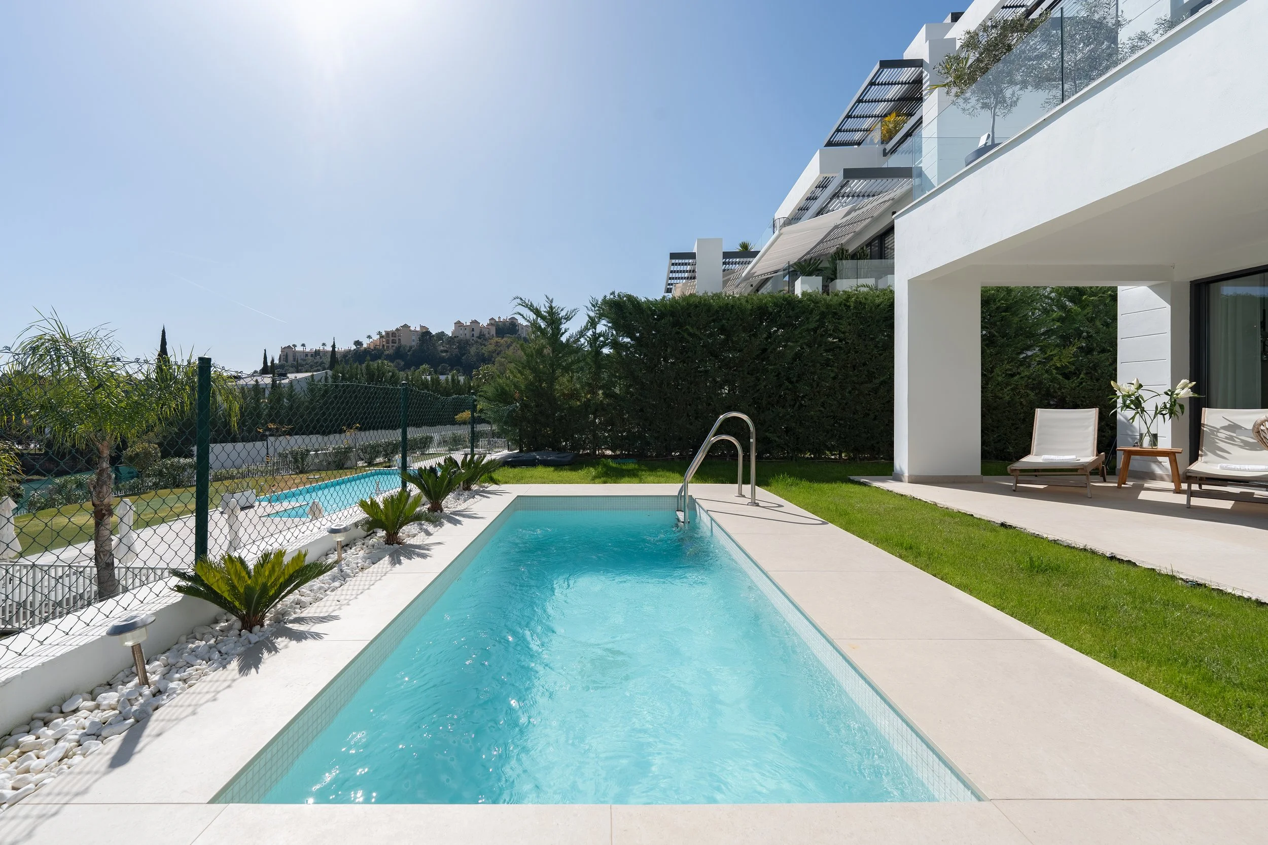 Modern backyard with a small swimming pool, white exterior walls, two lounge chairs, and potted plants under a covered patio. Green grass, trees, and a residential hillside in the background under a clear blue sky.