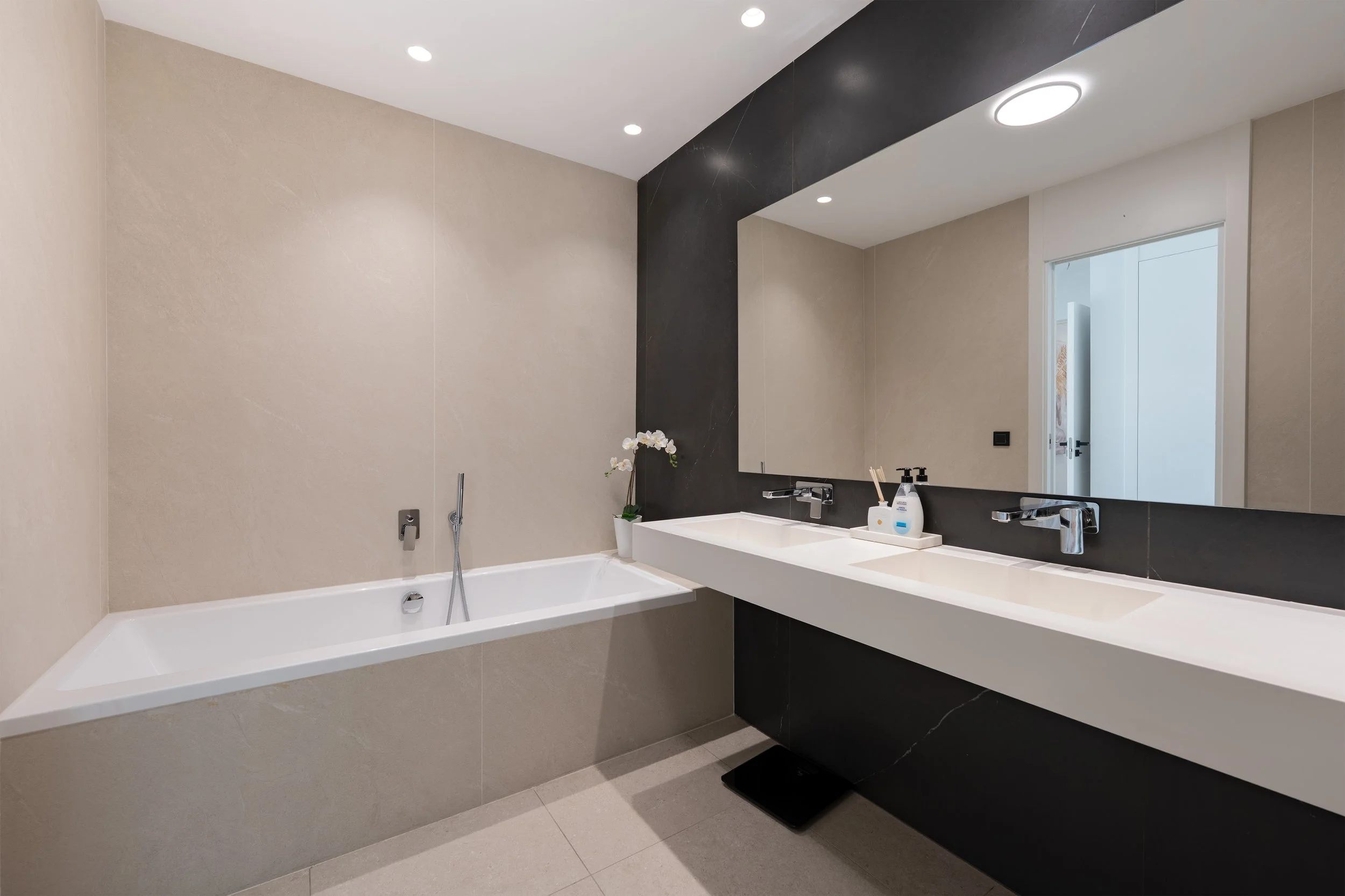 Modern bathroom with a built-in bathtub and a double sink vanity with large mirror, beige and black wall decor, and minimal accessories.