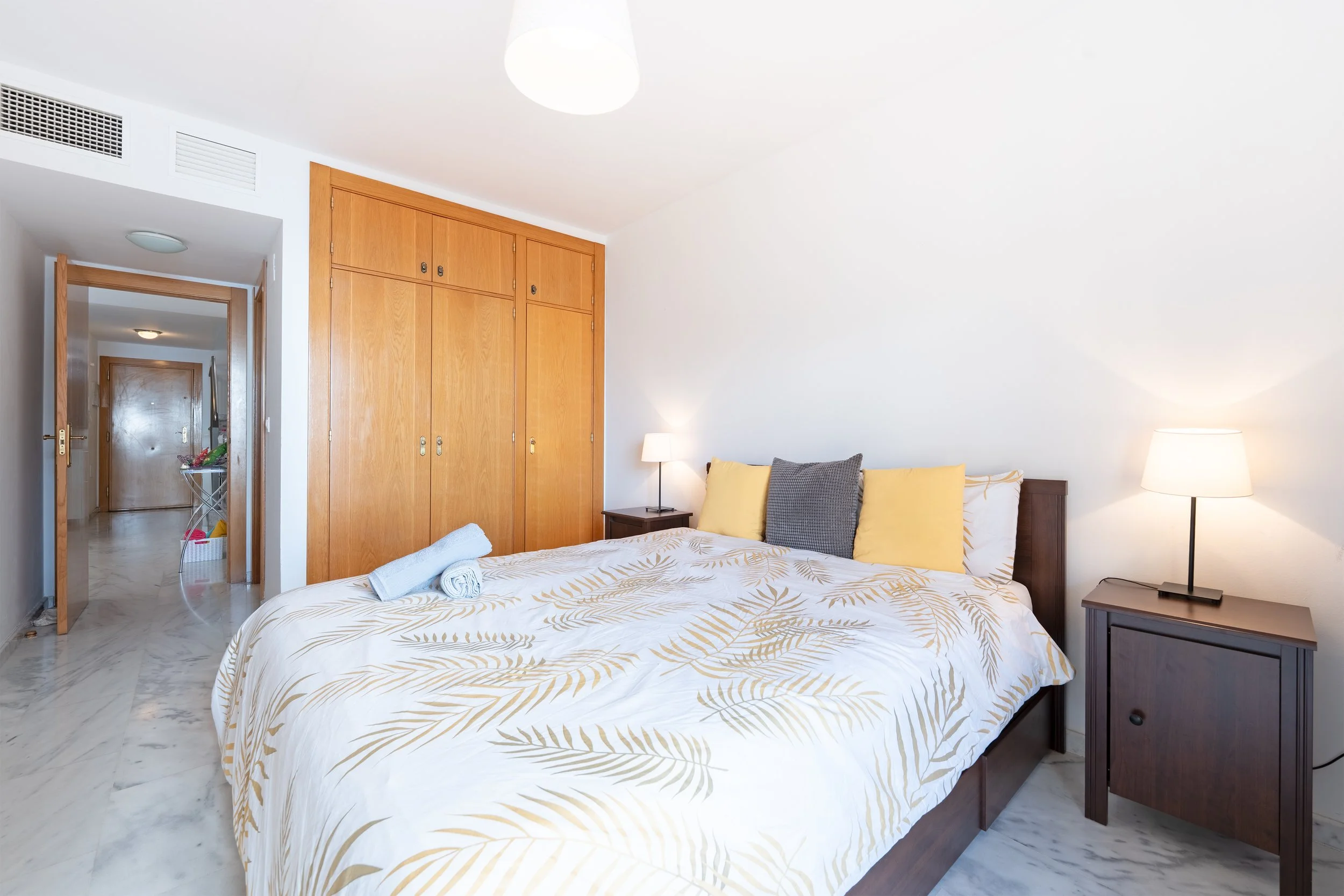 A bedroom with a bed featuring a white and gold leaf patterned duvet, yellow and gray pillows, a small bedside table with a lamp, a wooden wardrobe, and an open doorway showing a hallway with a laundry basket.