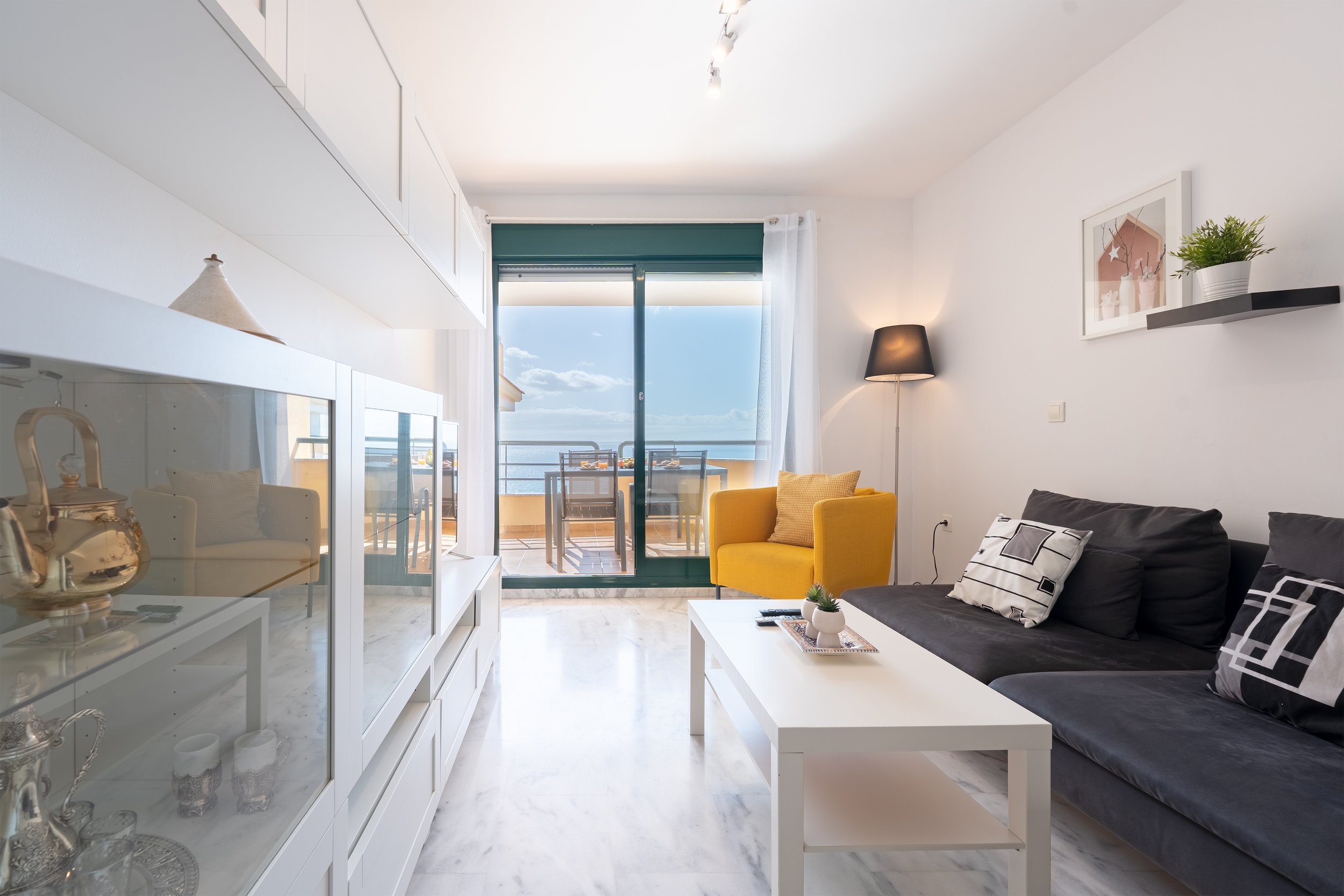 Living room with a sliding glass door leading to a balcony overlooking the sea, yellow armchair, black sofa with pillows, white coffee table, TV cabinet, and wall shelves with plants.