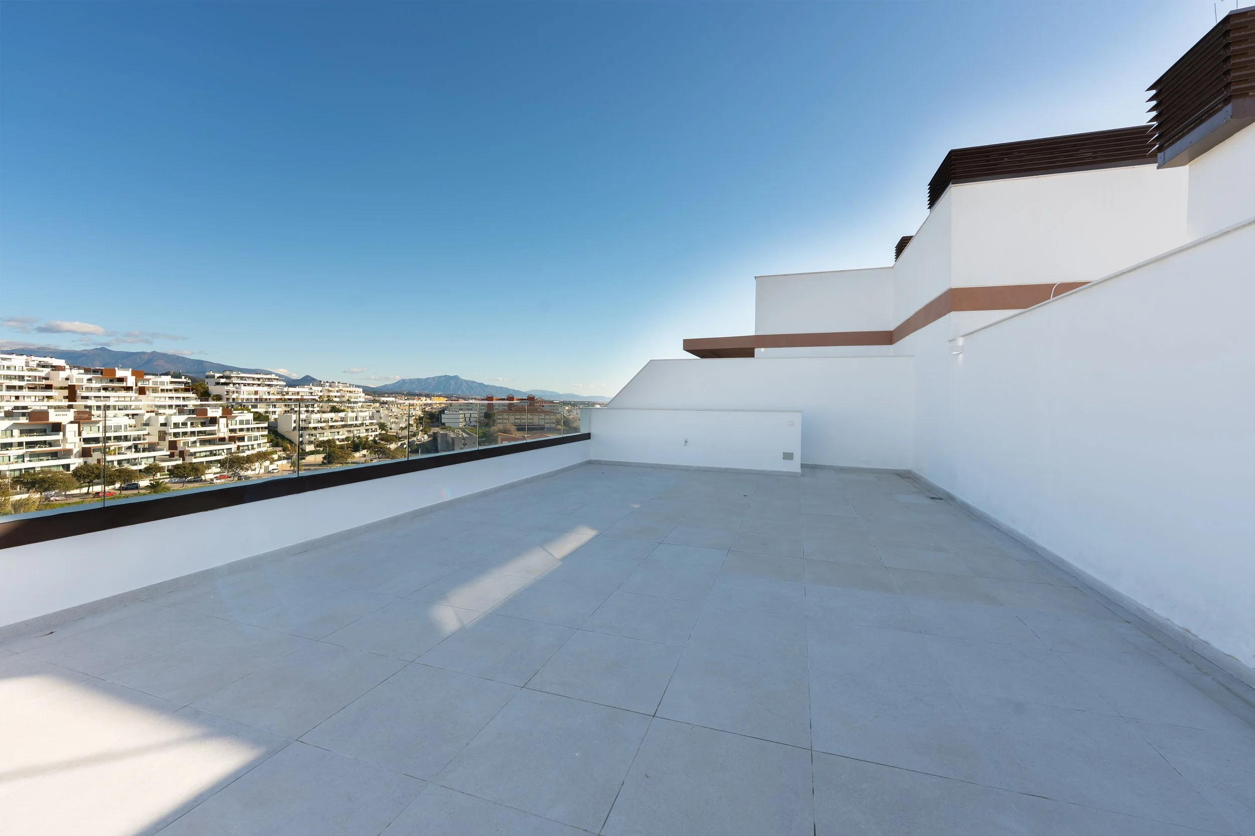 A spacious rooftop terrace with white walls, glass railing, and light grey tiled flooring, offering a view of residential apartment buildings and mountains in the distance under a clear blue sky.