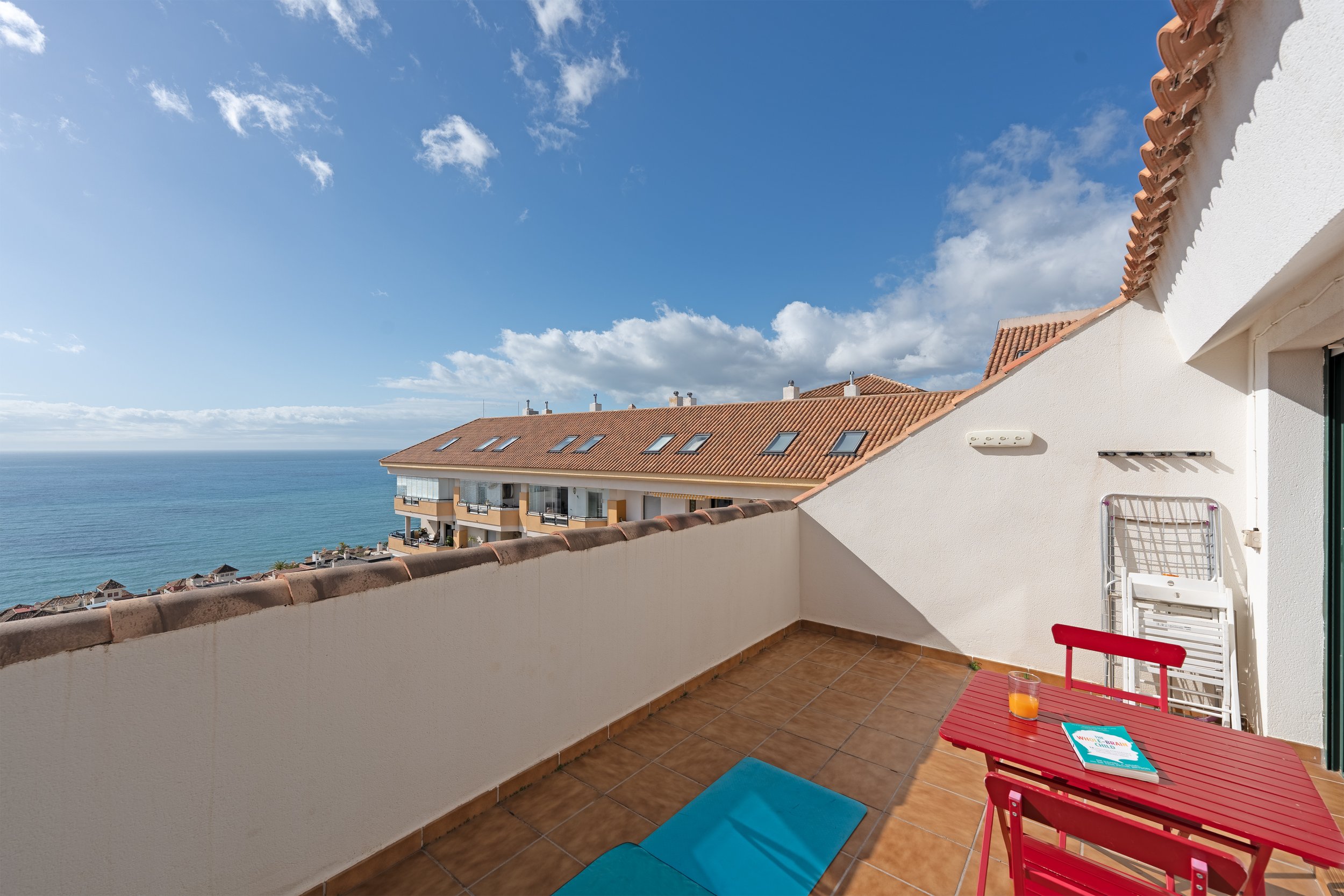 Balcony with terracotta tiles overlooking the ocean, with a red table, a glass of orange juice, a book, and outdoor furniture, with neighboring buildings and a partly cloudy sky.