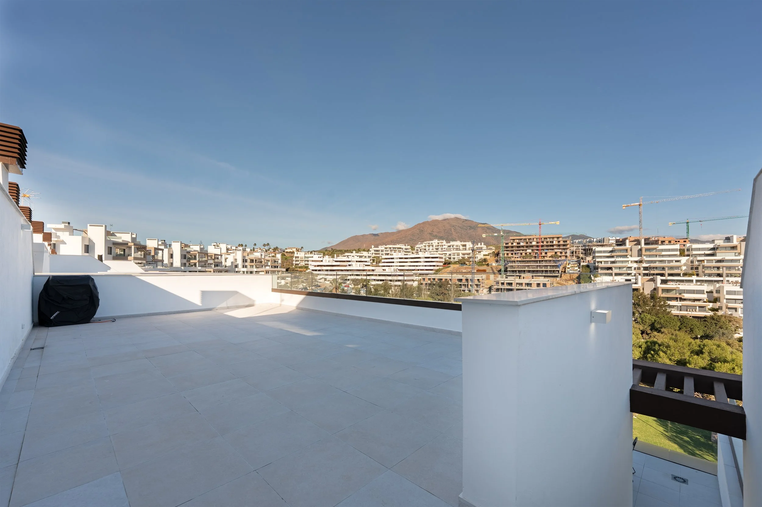 Empty balcony with white walls and tiled floor, overlooking a cityscape with modern white buildings under a clear blue sky and mountains in the background.