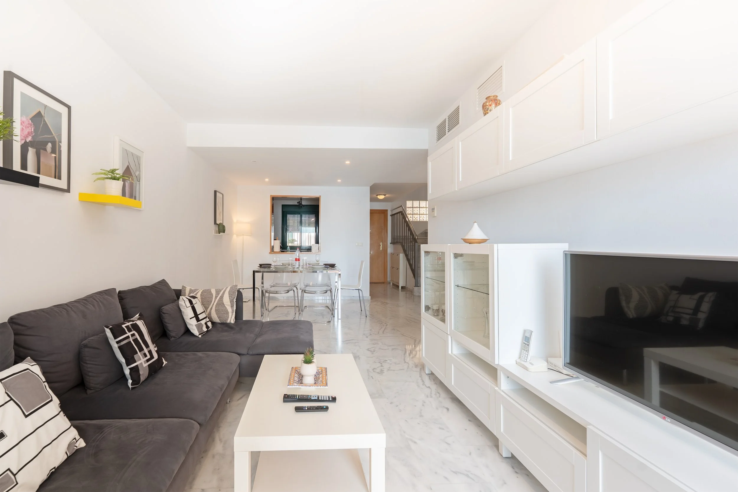 Living room with gray sectional sofa, black and white patterned pillows, white coffee table, flat-screen TV, white entertainment unit, dining area in background, artwork on walls, and marble flooring.