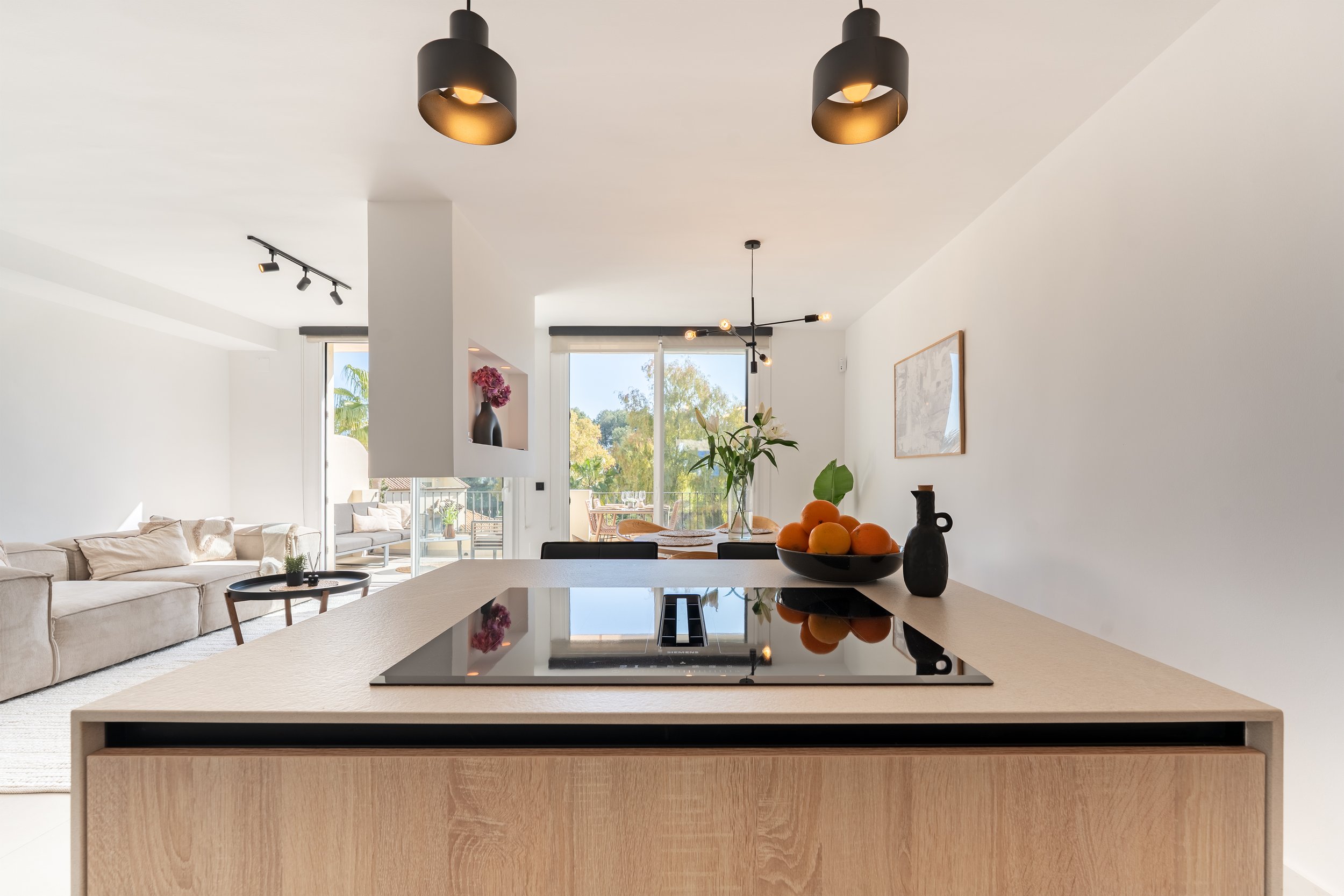 Modern open-concept living space with a kitchen island, black cooktop, black pendant lights, a beige sofa, artwork, and balcony doors showing outdoor greenery.