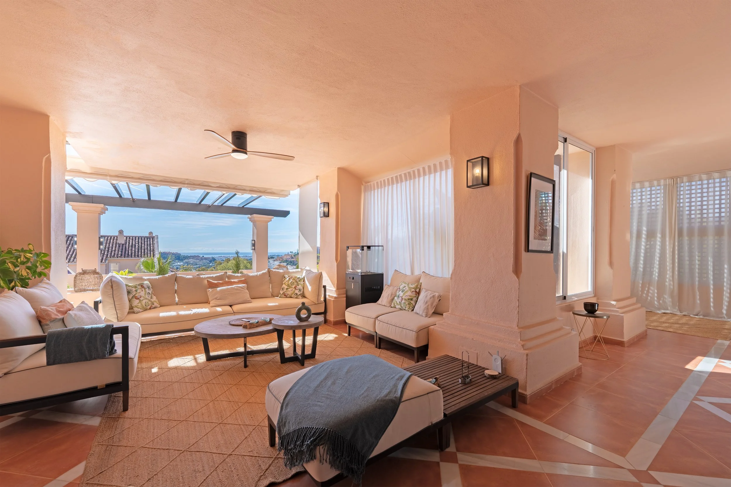 Bright and airy living room with cream-colored sofas, wooden coffee tables, and decorative pillows. Large open window with ocean view and sheer curtains, beige tiled floor with intricate border design, ceiling fan, and wall-mounted light fixtures. De