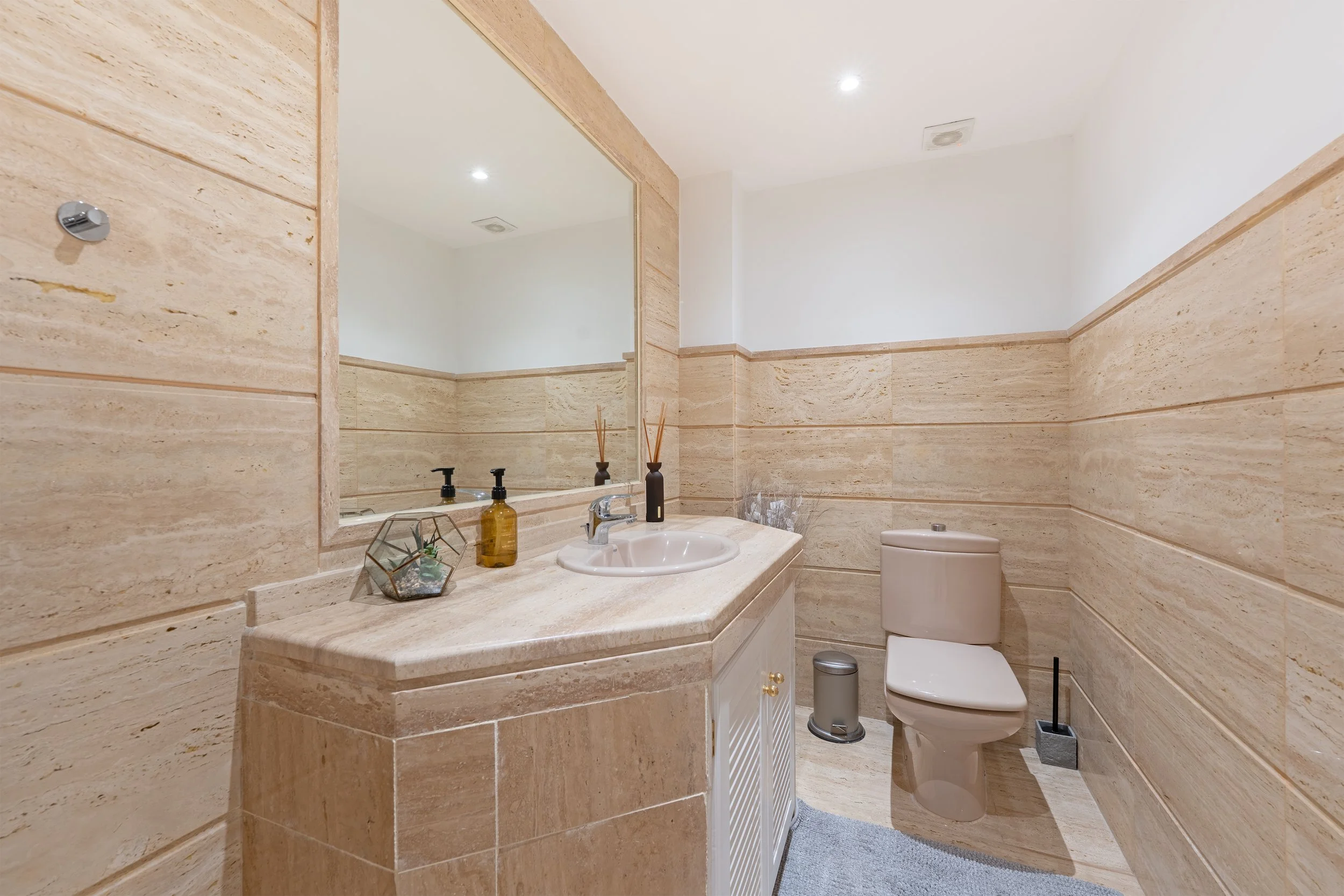 A beige bathroom with a large mirror, a sink with a marble countertop, soap dispenser, decorative items, and a toilet, with wood-like tiles on the walls and floor.