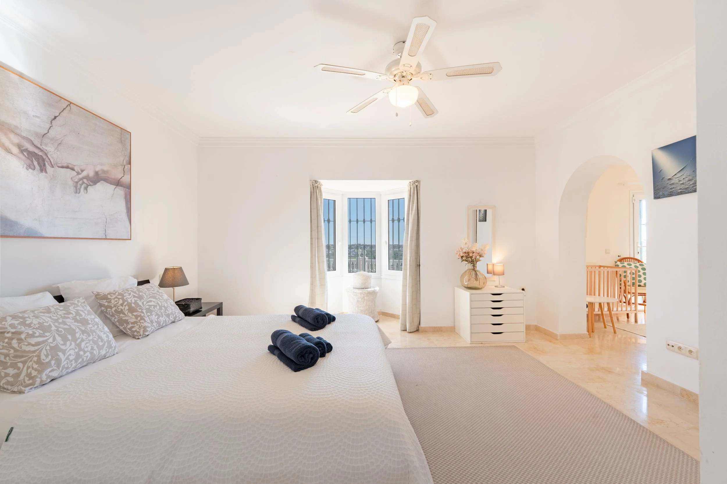 Bright, airy bedroom with a large bed decorated with patterned pillows and rolled towels, a nightstand with a lamp, a dresser with a vase and flowers, a window with curtains, and a ceiling fan.
