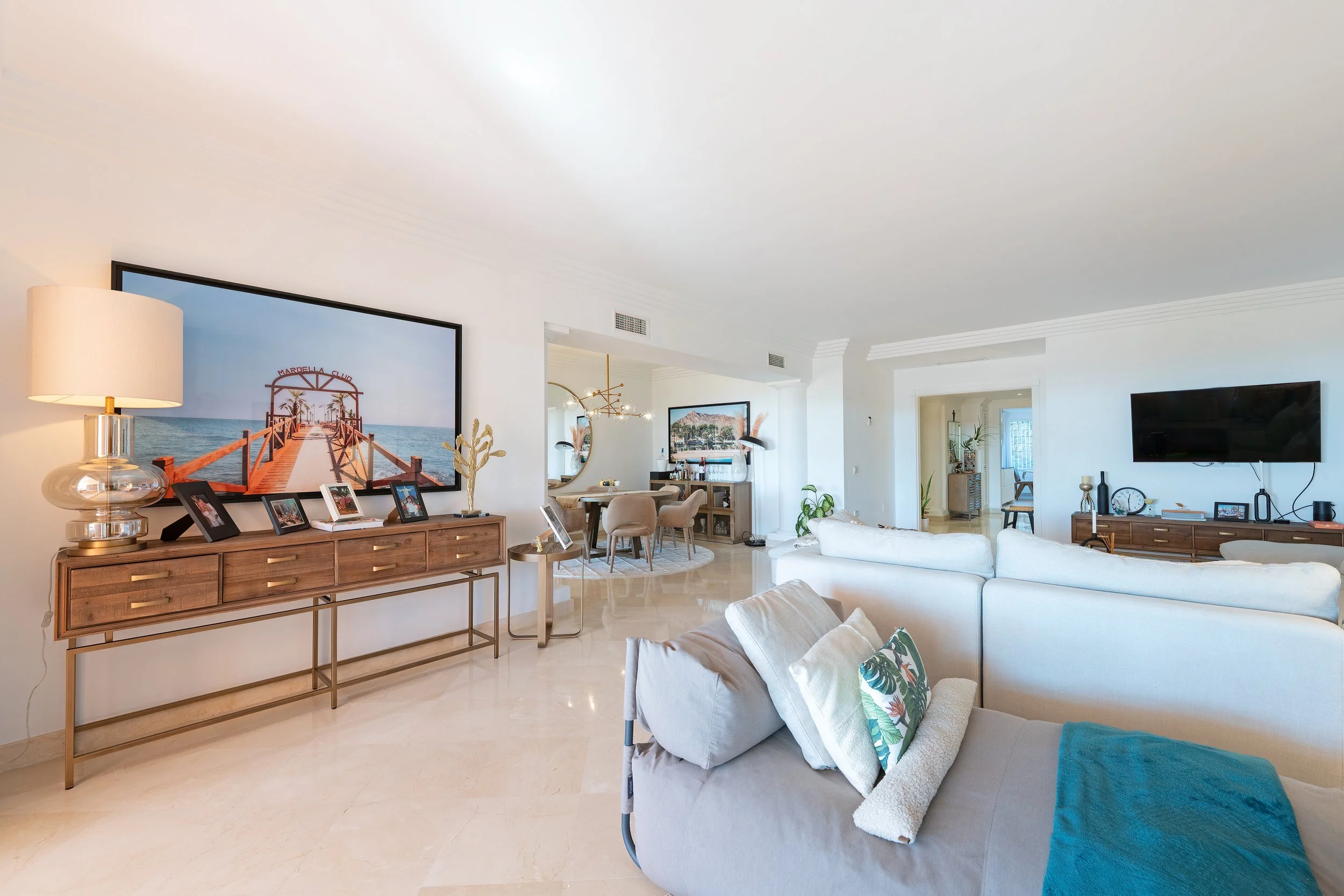 Spacious living room with white walls and flooring, featuring a large wall-mounted flat-screen TV, a white sofa with patterned pillows, a wooden console with photo frames and decorations, a round dining table with chairs, and various plants and decor
