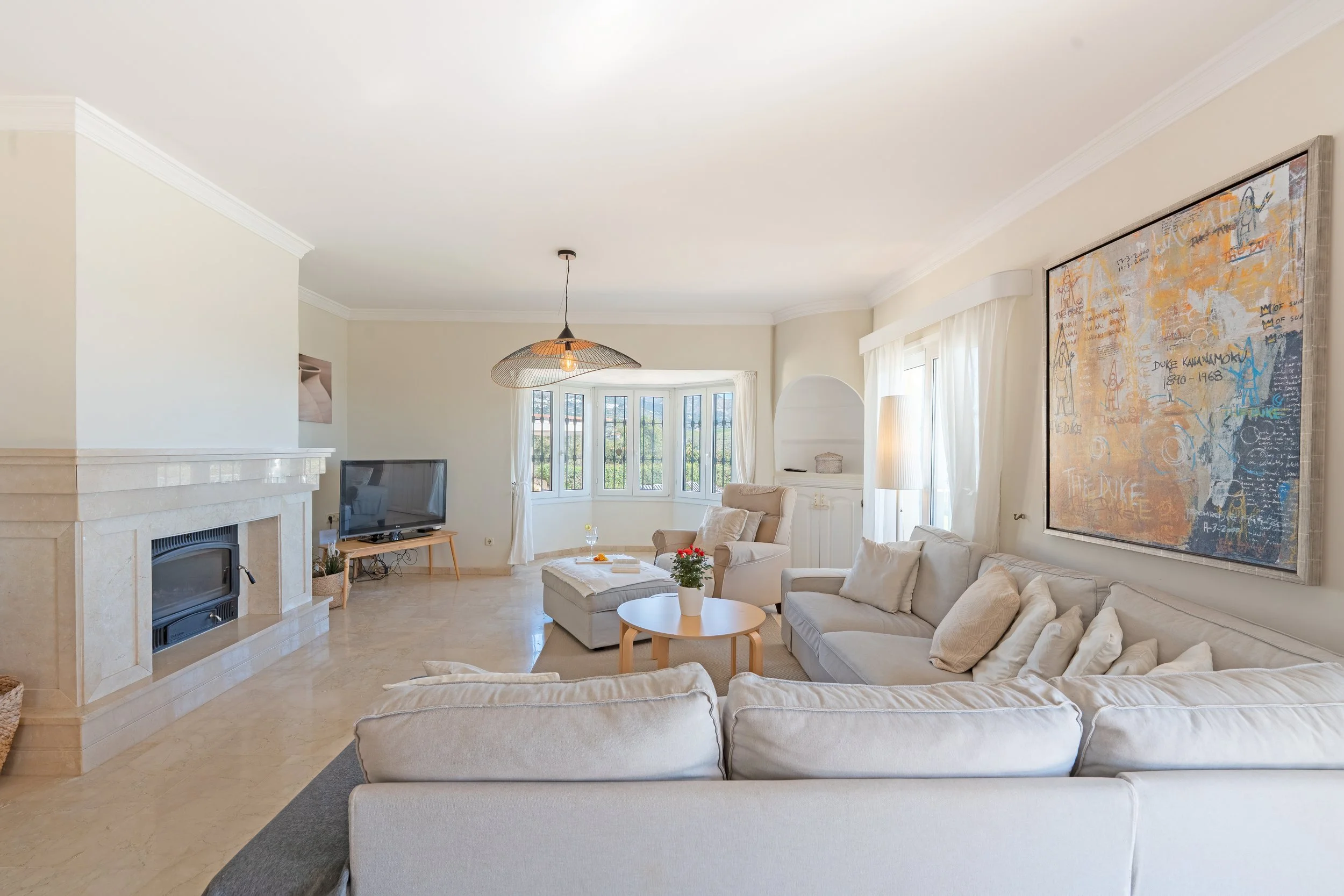 A bright, spacious living room with cream-colored sofas, a wooden coffee table with a plant, a TV on a stand, a fireplace, large windows, and a large colorful abstract painting on the wall.