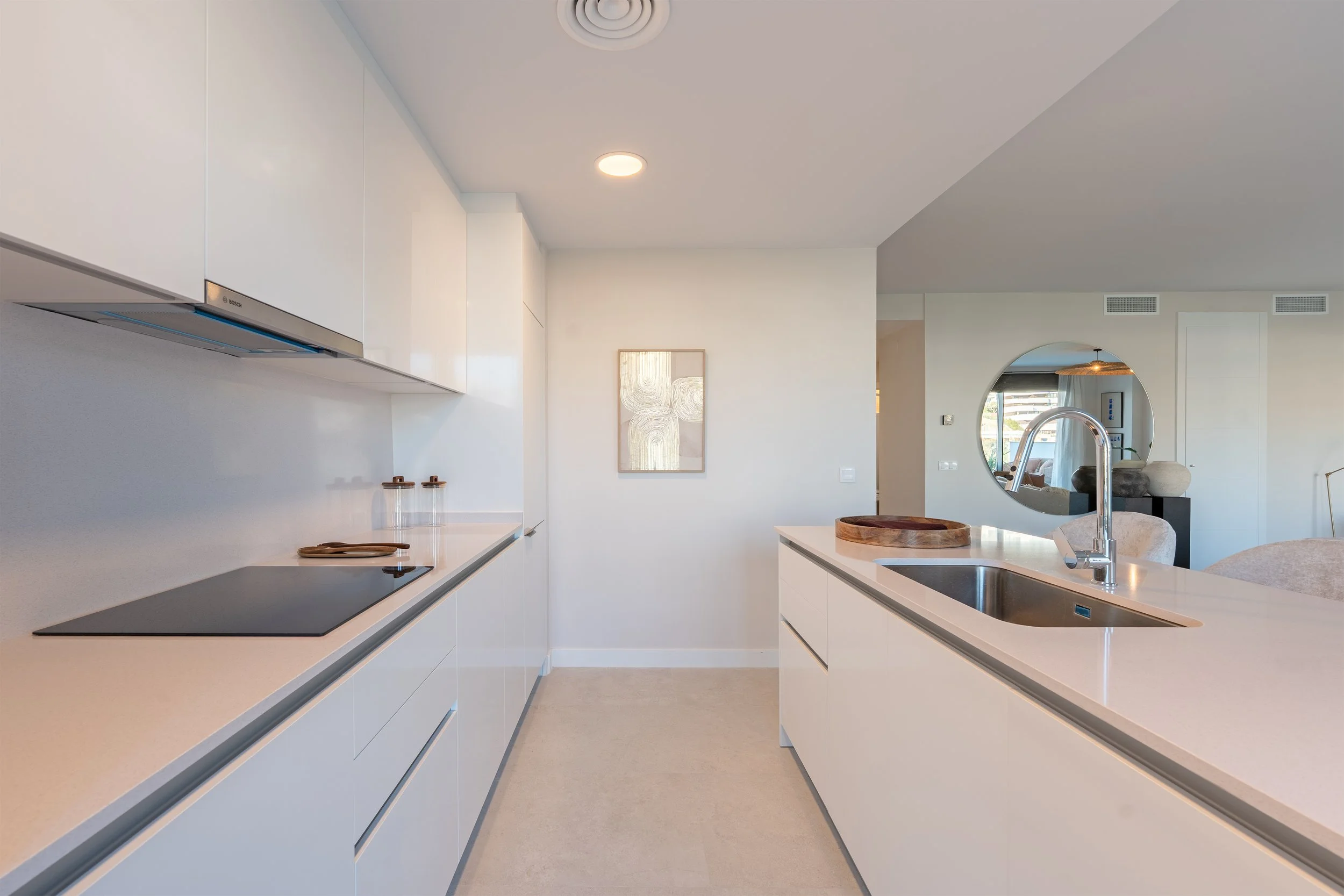 Modern kitchen with white cabinets, beige countertops, a stove, a sink with a chrome faucet, and a circular mirror on the wall reflecting an adjacent room.