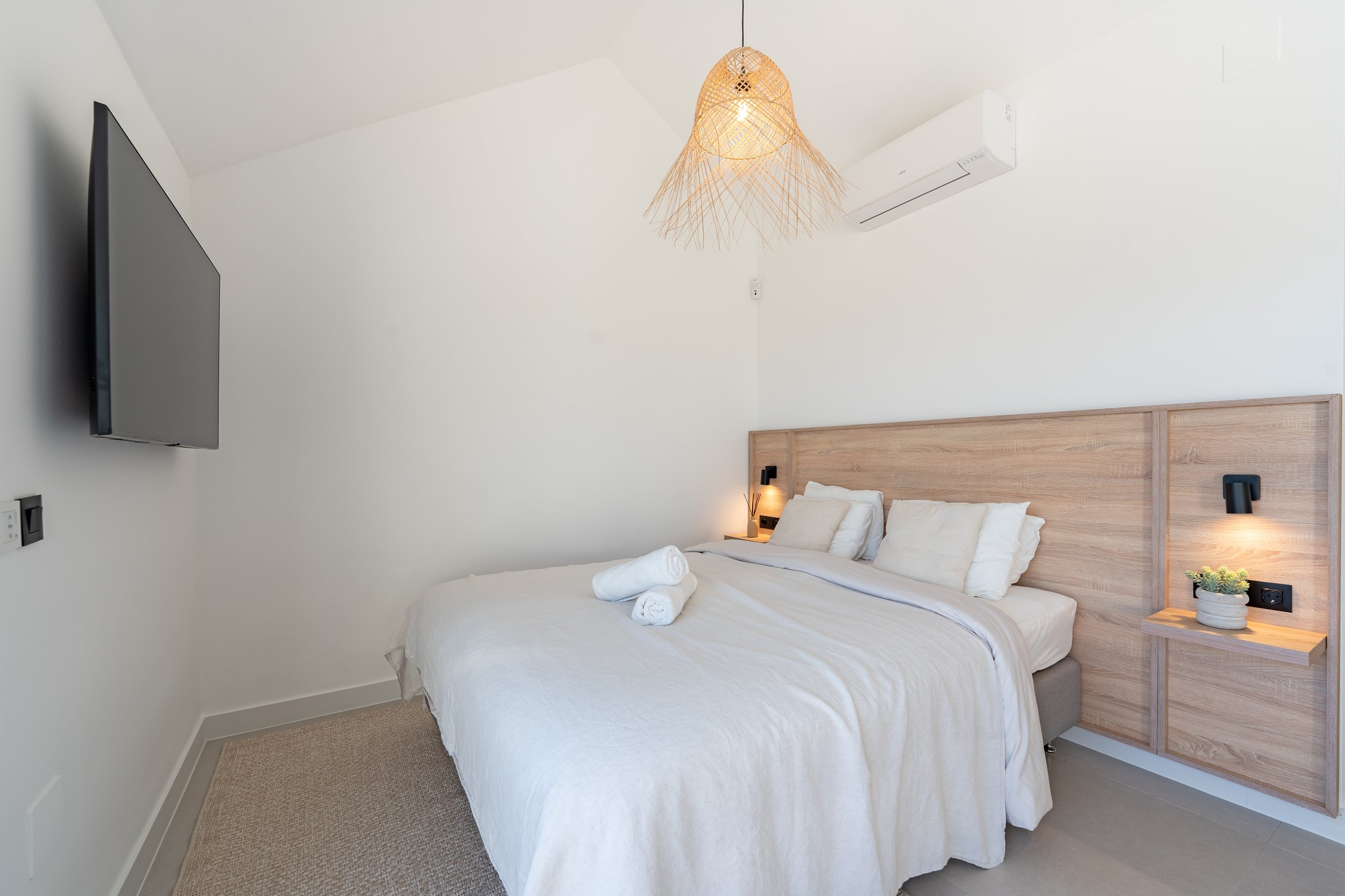 A neatly made bed with white linens and pillows, two rolled towels on top, in a minimalistic bedroom with white walls, a wooden headboard, wall-mounted black reading lights, a small wooden shelf with a potted plant, a wall-mounted television, an air 