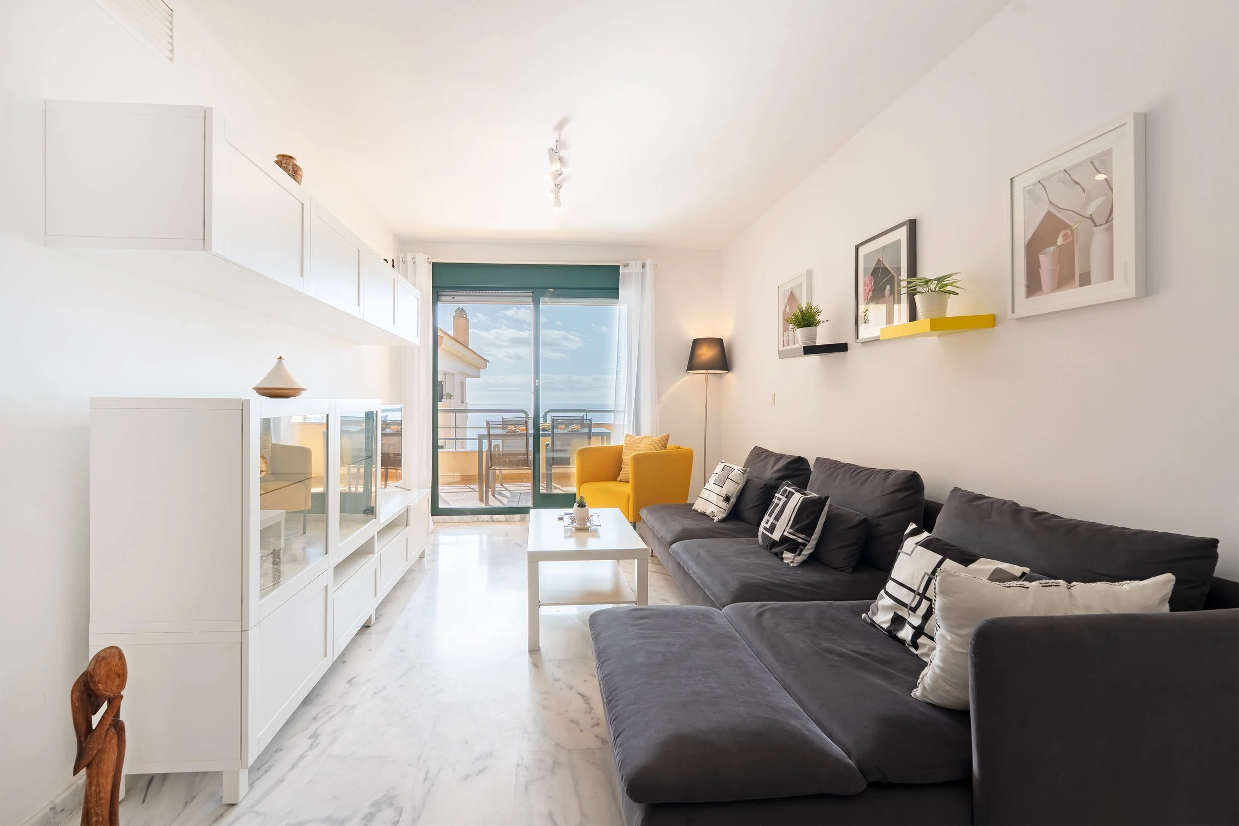 A modern living room with a large dark gray sofa, yellow accent chair, white coffee table, and decorative framed artwork on the wall. There is sliding glass door leading to a balcony with outdoor furniture, and sunlight coming in through sheer curtai