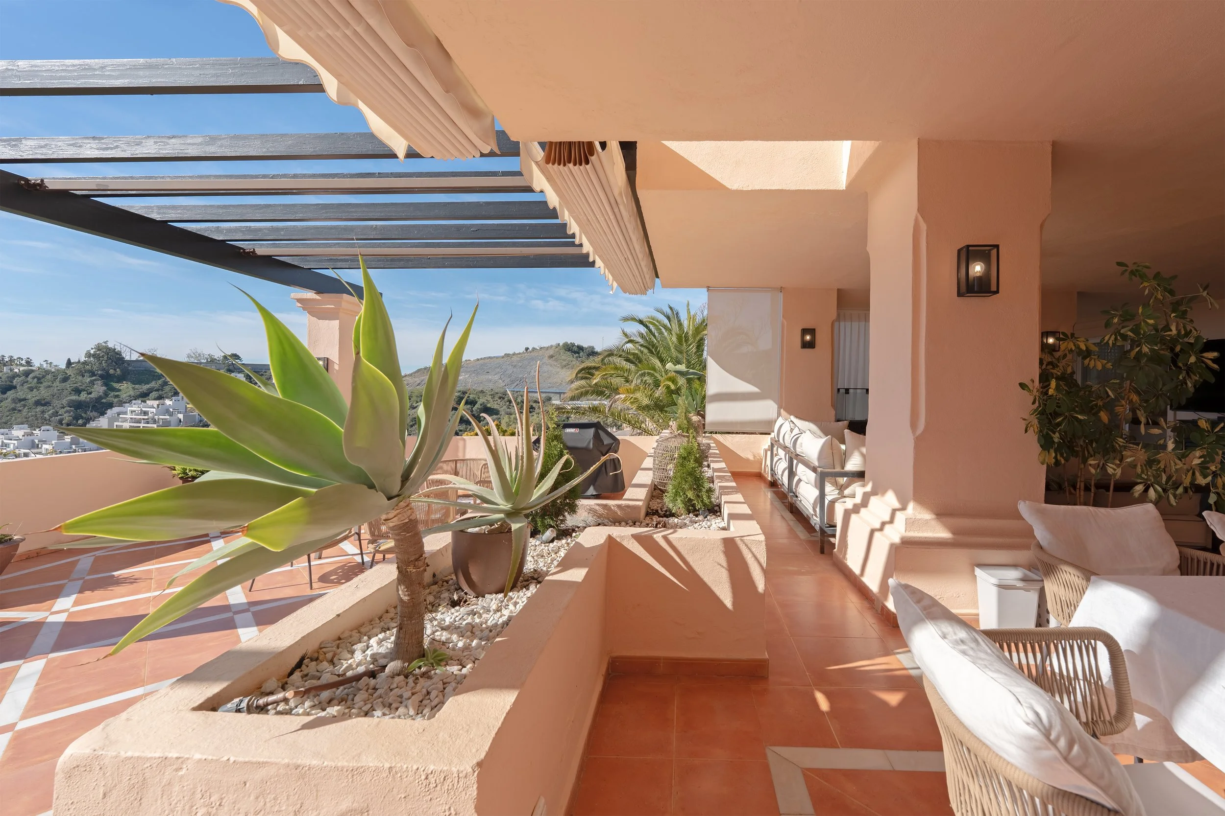 Outdoor terrace with potted succulents, white and beige furniture, palm trees, and panoramic view of hills under a blue sky.