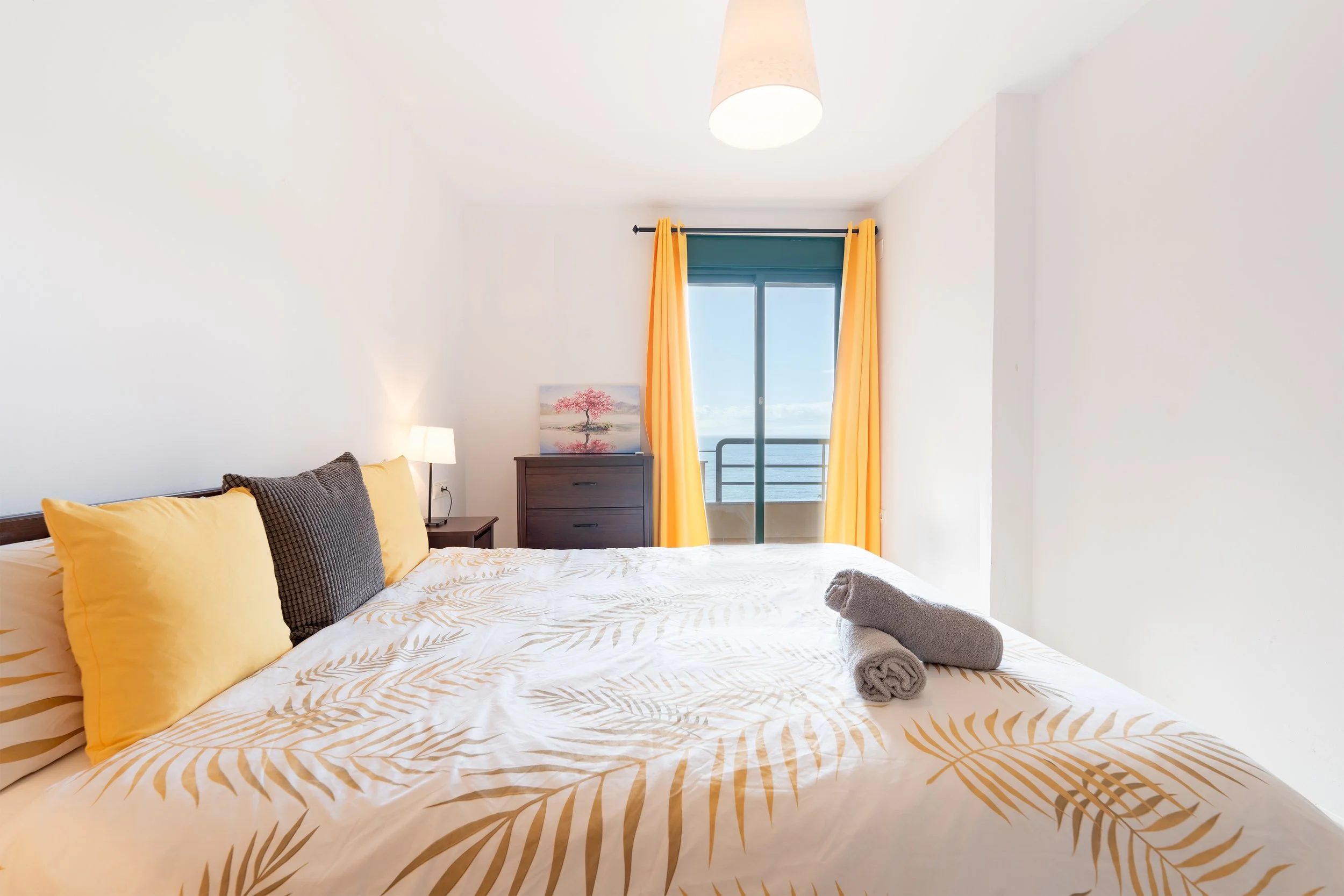 Bedroom with a bed, yellow pillows, gray towel, nightstand, lamp, chest of drawers, painting, and balcony with ocean view