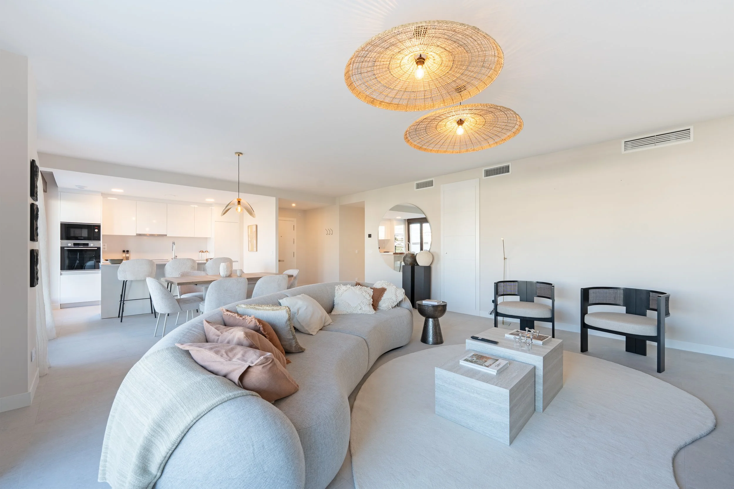 Modern living room with a large curved sofa, cream-colored pillows, two black circular chairs, and natural light from windows. Light wood coffee table, textured rug, and decorative lighting fixtures on the ceiling.