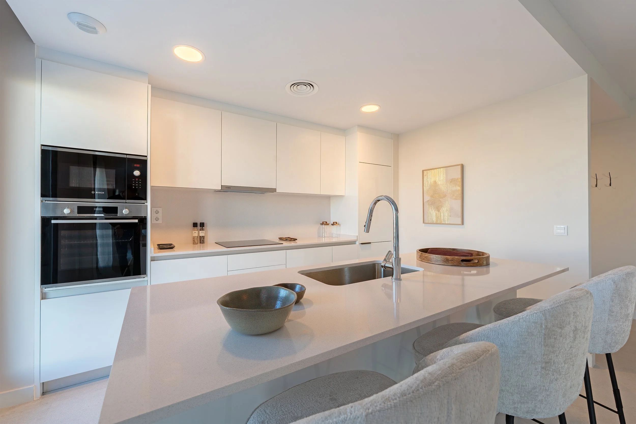 Modern kitchen with white cabinets, a built-in oven and microwave, a white countertop island with a sink, bowls, and a tray, beige chairs, and minimalist decor.