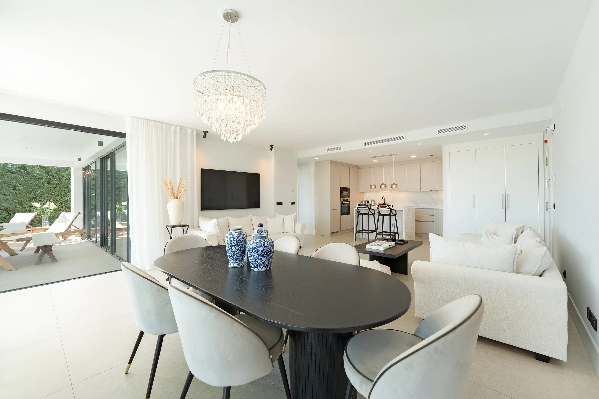 Modern open-concept living room and kitchen with white furniture, black dining table, blue and white vases, large window, and outdoor patio area