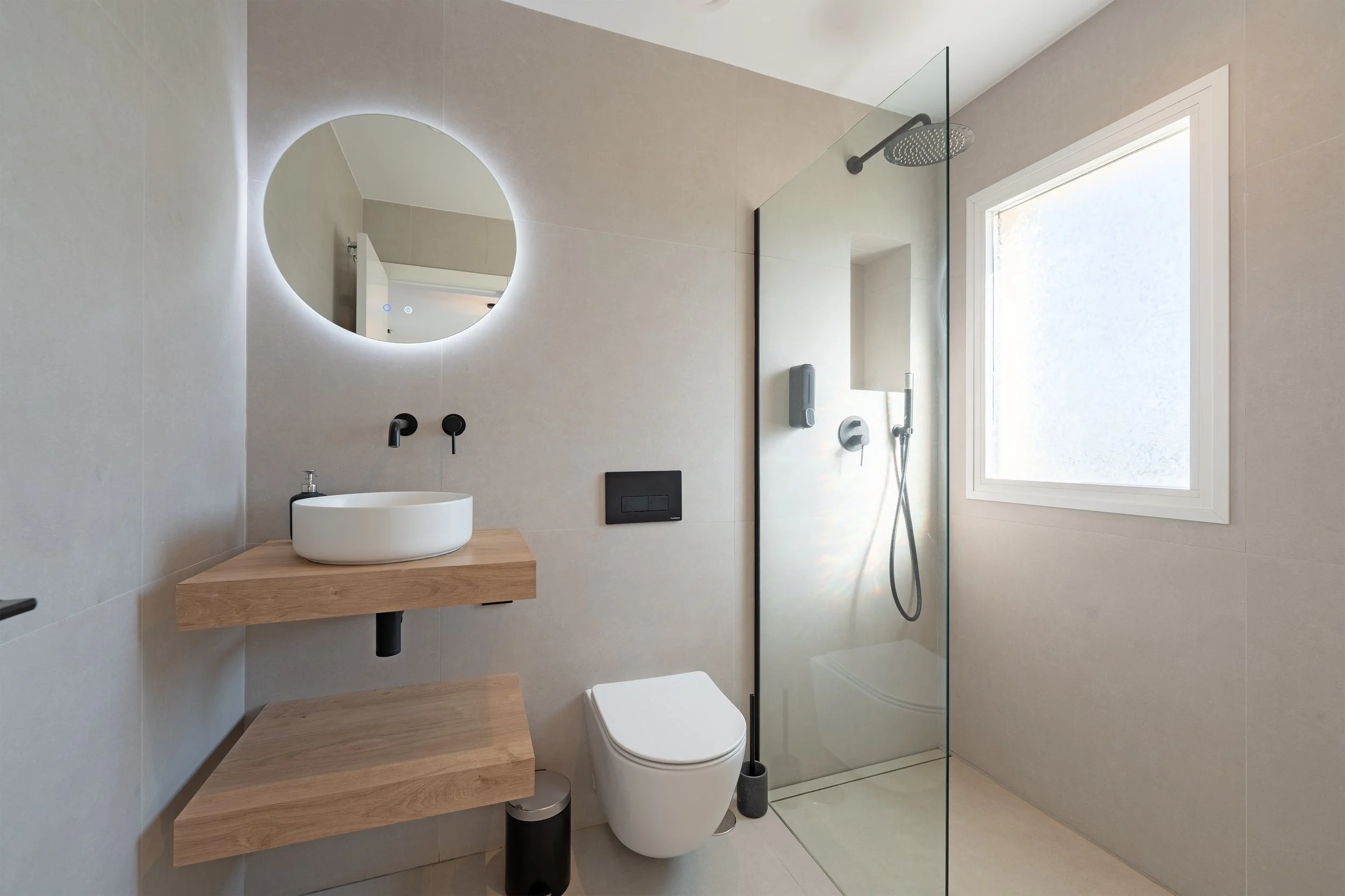 Modern minimalist bathroom with neutral color scheme, circular illuminated mirror, small vessel sink on wooden countertop, wall-mounted toilet, and walk-in shower with frosted window and rainfall showerhead.