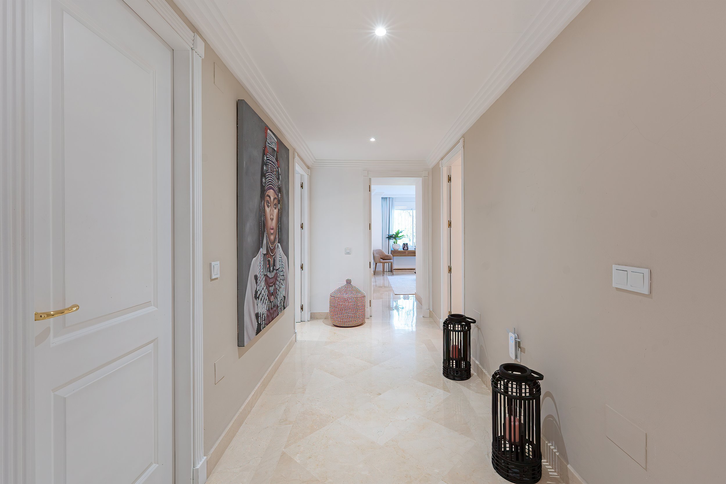 Empty hallway with beige walls, white trim, and tiled floor leading to a bright room with window, blue curtains, and a chair. Art piece of a woman in traditional attire on the left wall. Two black decorative candle lanterns on the right, near a white