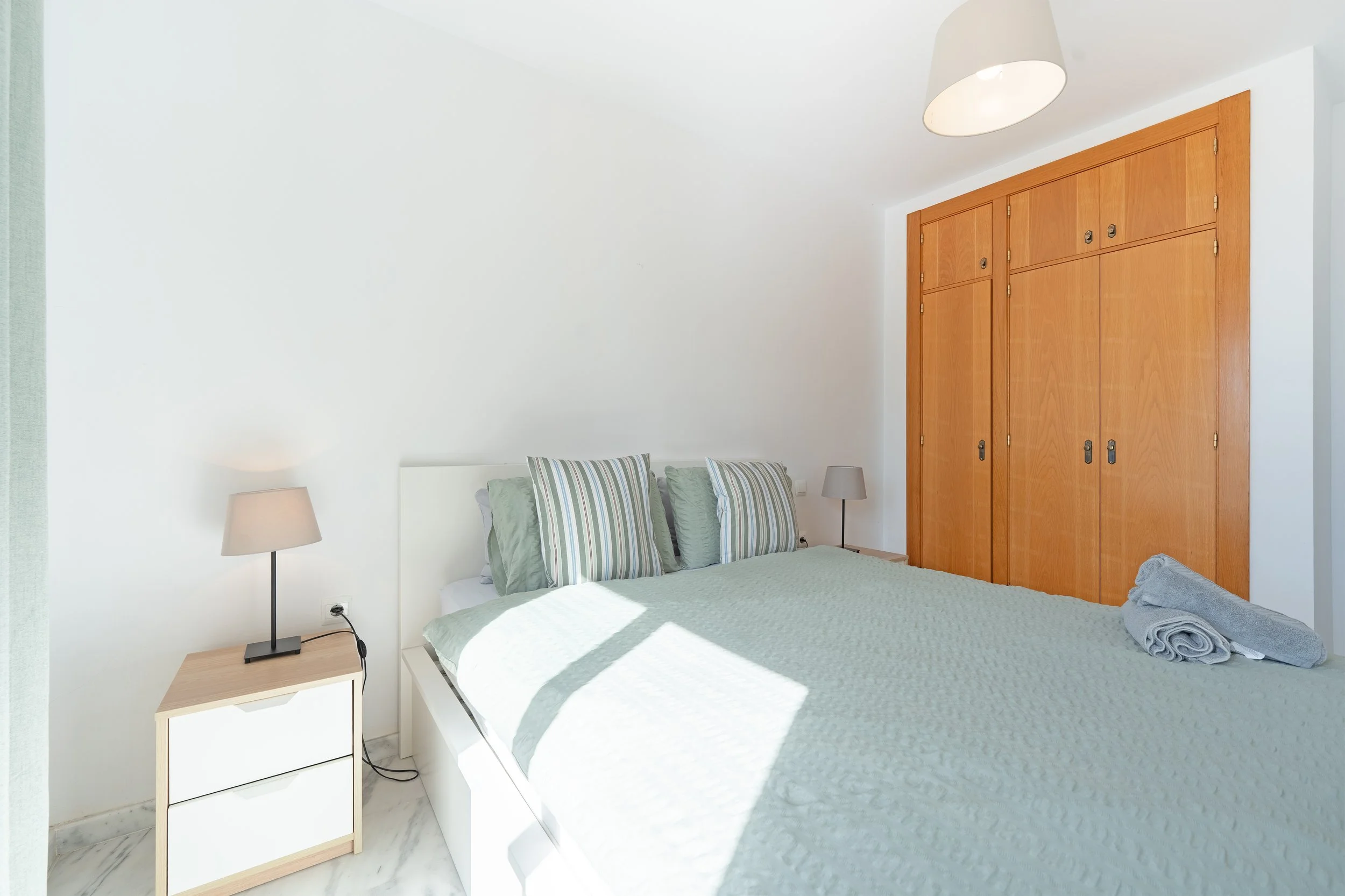 A bedroom with a white bed, striped pillows, two nightstands with lamps, a wooden closet, and a rolled towel on the bed.
