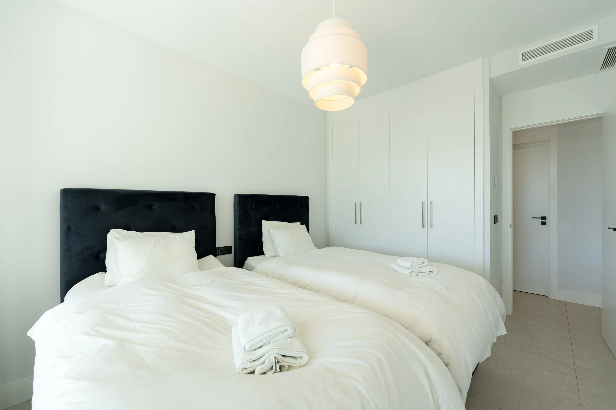 A modern, minimalist hotel room with two beds and black headboards, white bedding, folded towels on each bed, white walls, a white closet, and a ceiling light fixture.