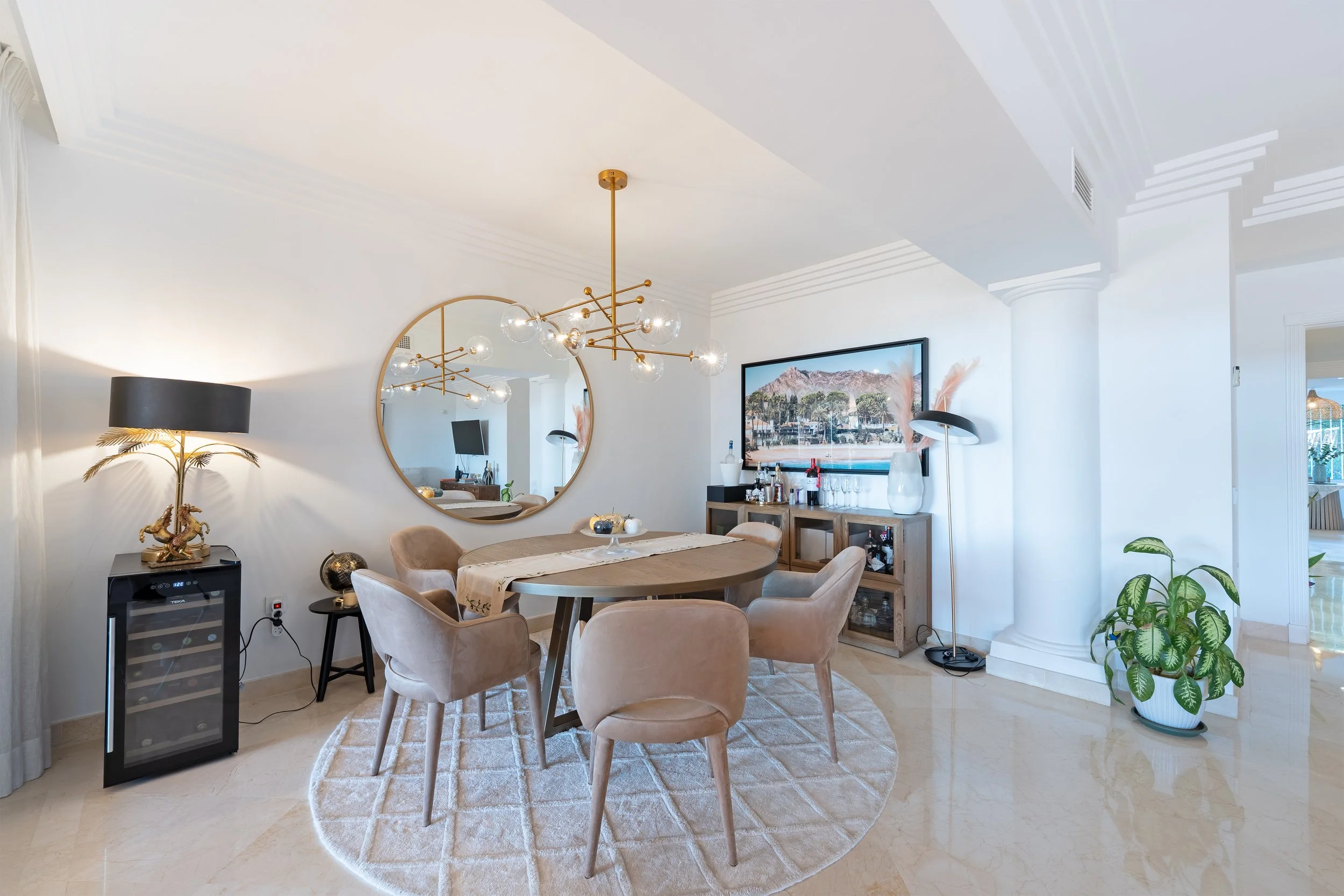 Living room with a wooden dining table surrounded by six beige upholstered chairs, a large round mirror on the wall, a modern chandelier above, a TV, a bar cabinet with drinks, a vase, and a floor lamp, with a potted plant in the corner.
