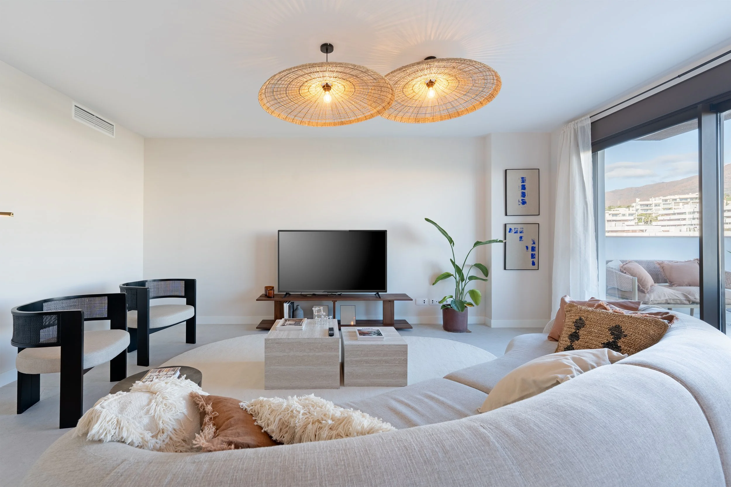 Modern living room with a large white sectional sofa, a flat-screen TV on a wooden stand, two beige stone coffee tables, two black chairs with white cushions, a large potted plant, and a sliding glass door with white curtains leading to an outdoor ba