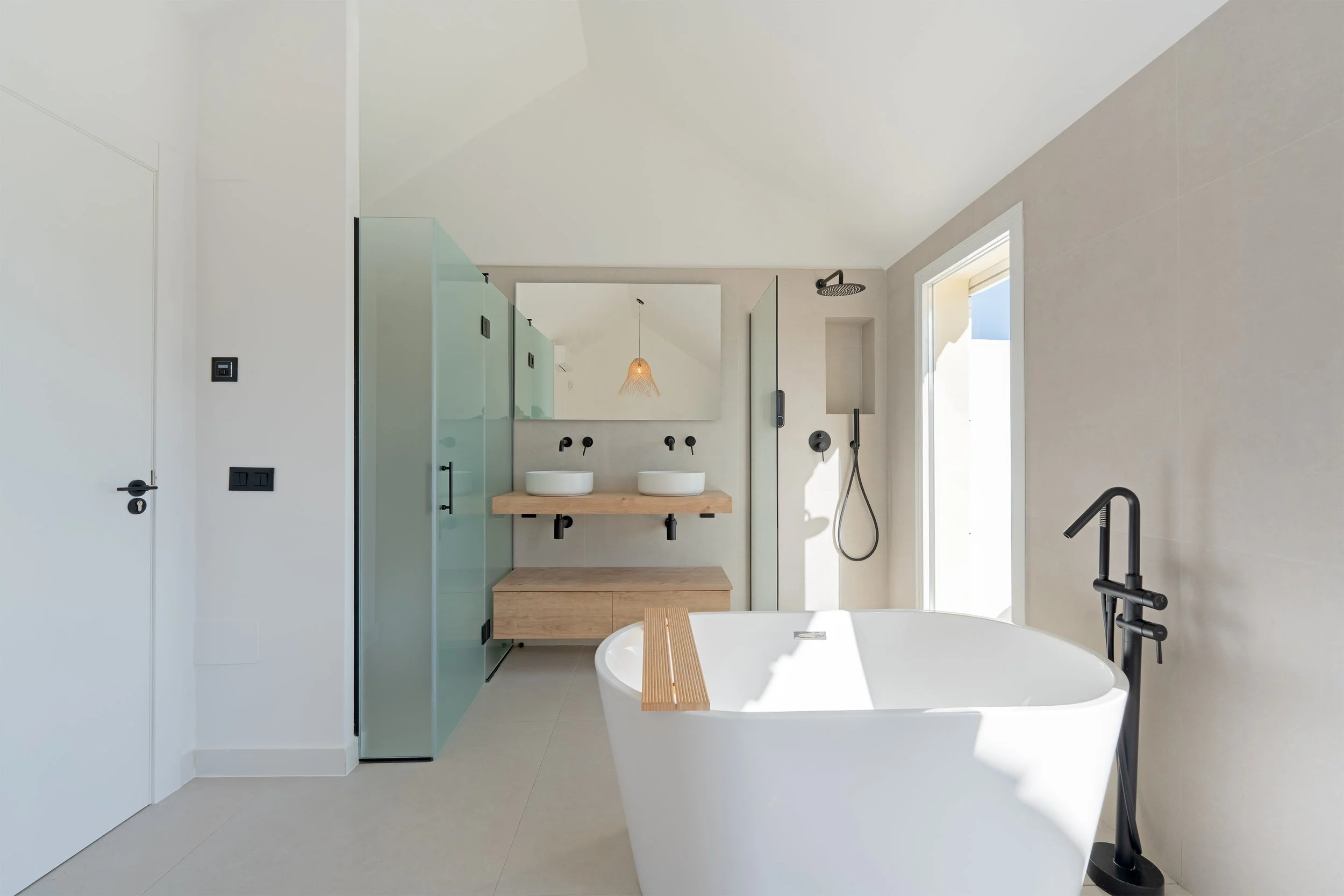 Modern bathroom with a freestanding bathtub, double sinks with wooden countertop, large mirror, walk-in shower with black fixtures, and a glass door leading outside, all in a minimalist style.