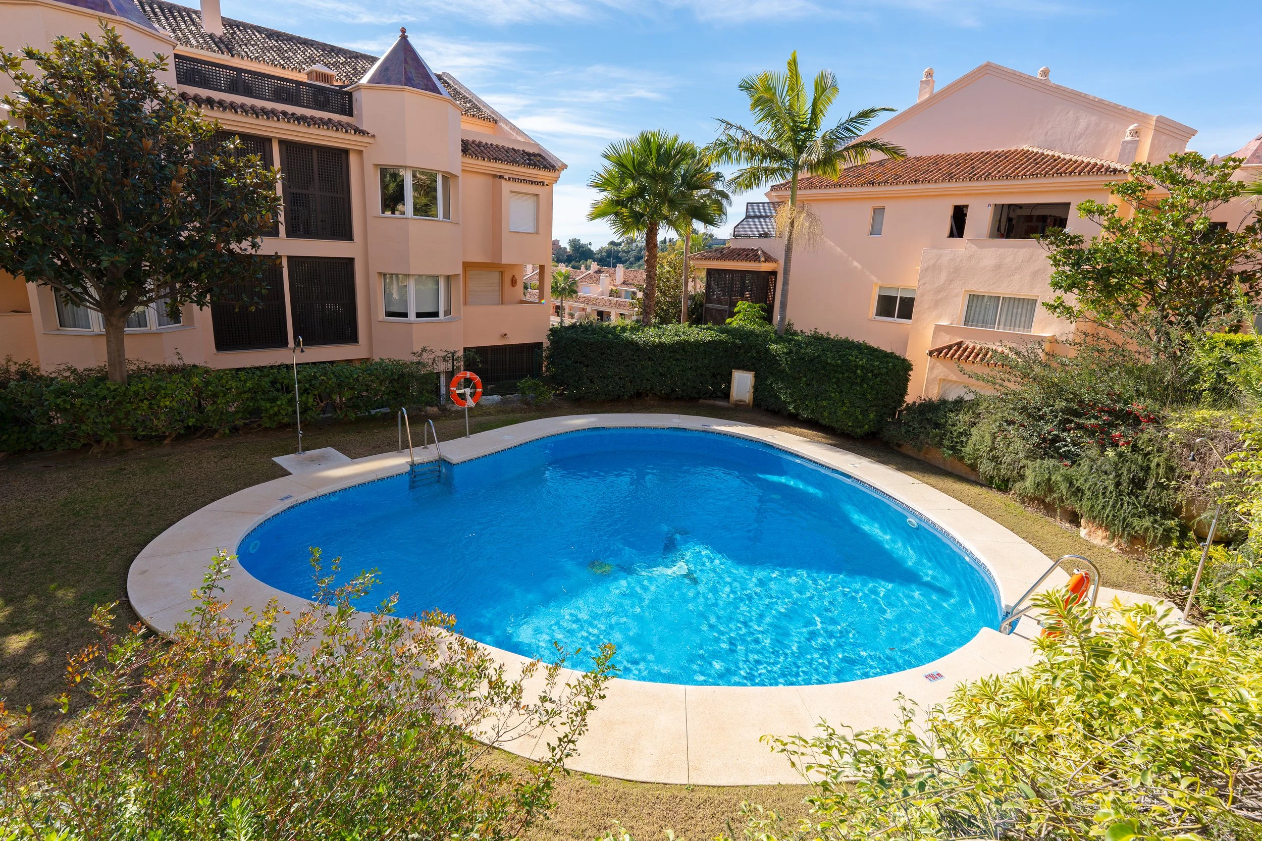 A swimming pool surrounded by a concrete deck in a residential complex with pink stucco buildings, palm trees, and landscaping.