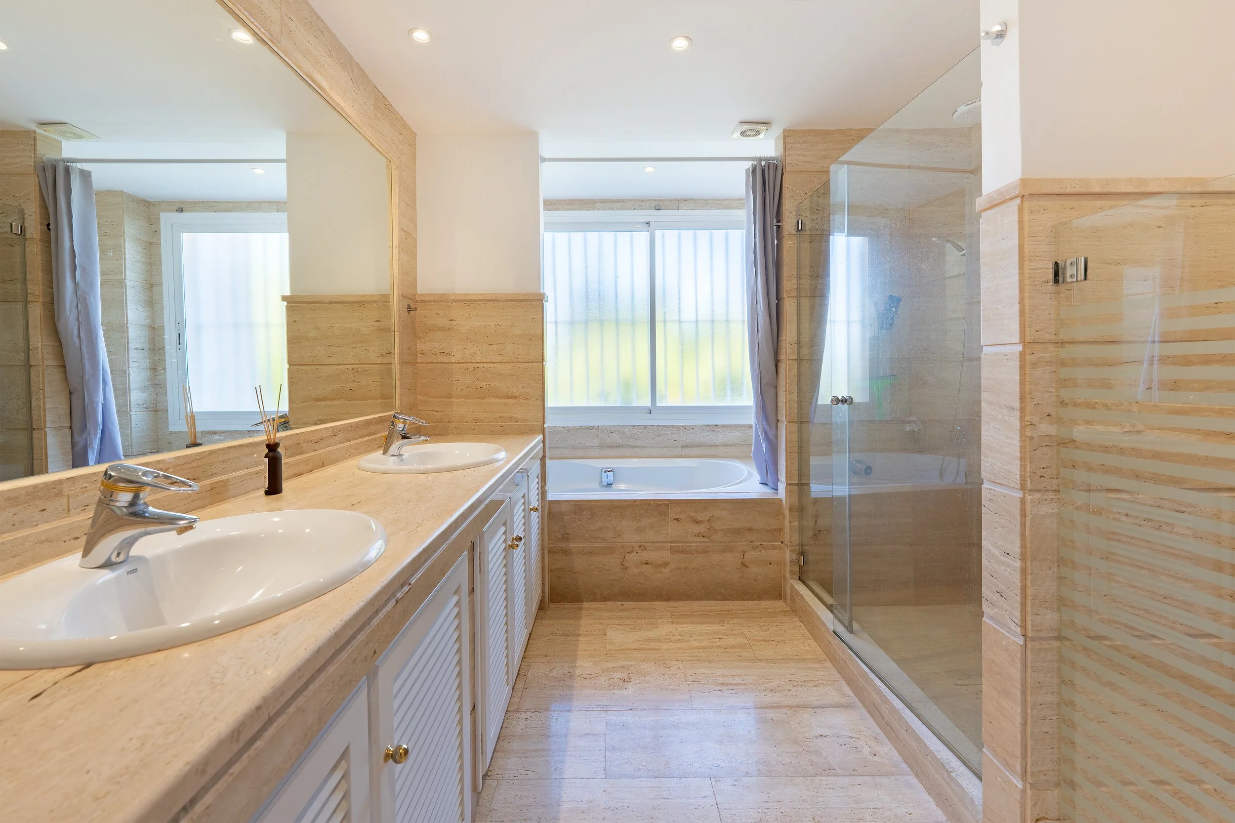 Bathroom with double sink vanity, large mirror, bathtub under a window, and glass-enclosed shower with beige tile decor.
