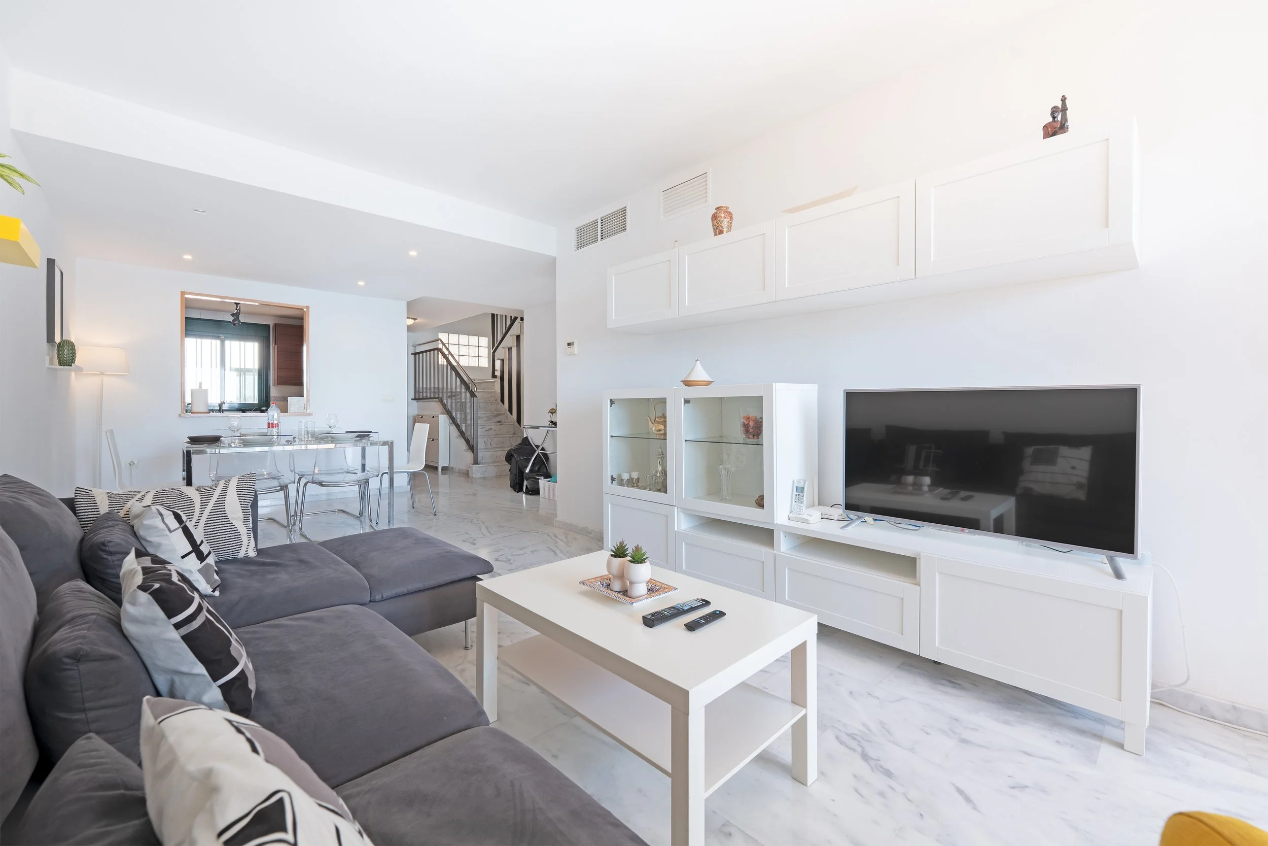Bright living room with white walls, marble floor, grey sectional sofa with black and white pillows, white TV stand with a large flat-screen TV, white coffee table with two remote controls and small potted plants, dining area with glass table and whi