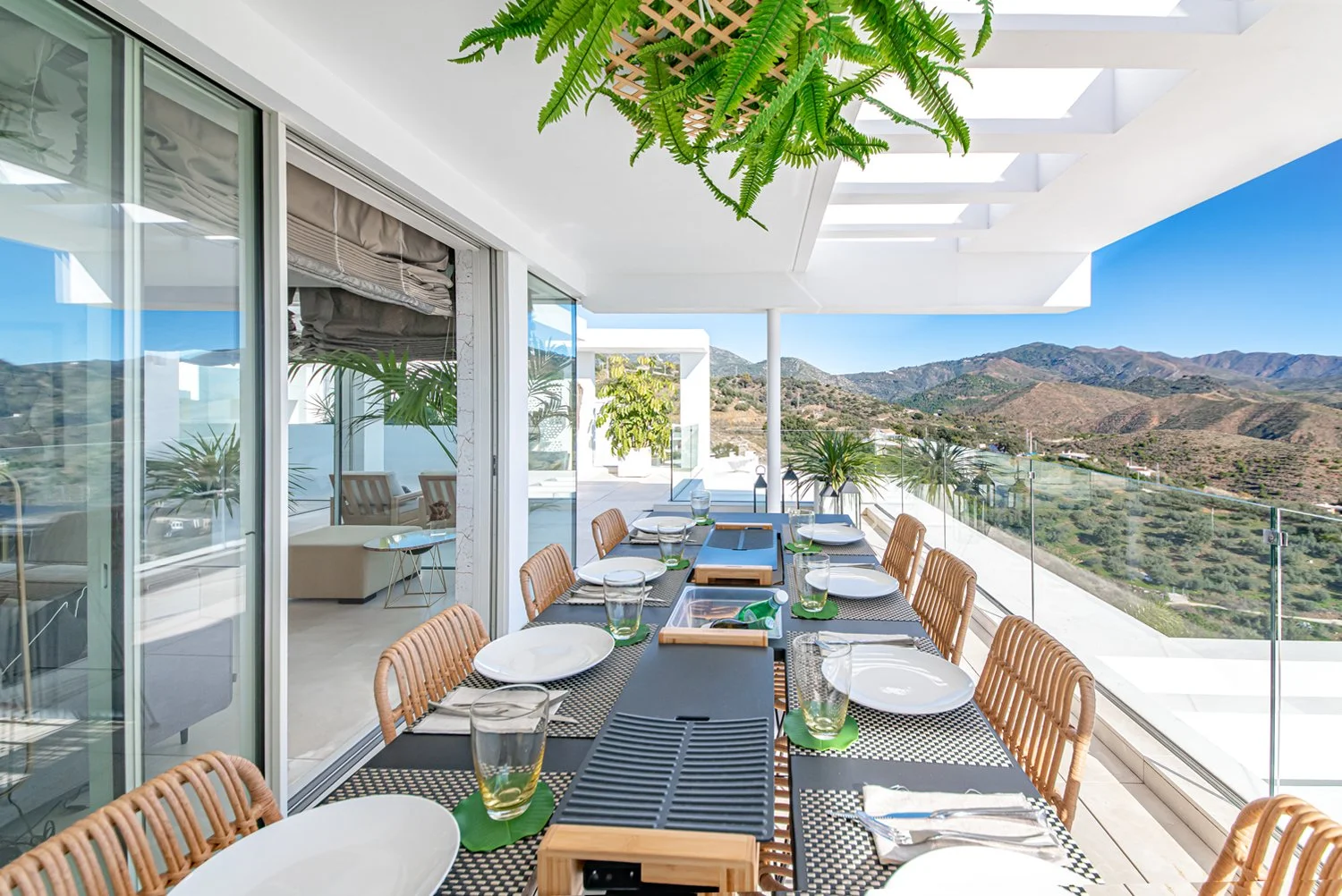 An outdoor dining area on a balcony with a long table set with white plates, glasses, silverware, and cutting boards. The balcony has glass railing and offers a view of rolling hills and mountains under a clear blue sky. Potted plants and a hanging f